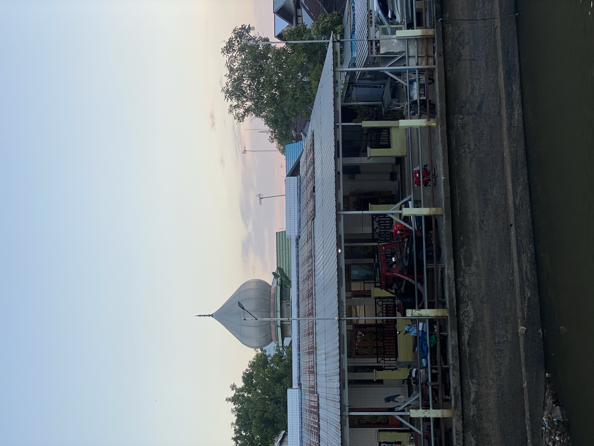 The image depicts a waterside scene in Tunggul Irang Ulu, Indonesia, featuring a building with a distinctive dome, likely a mosque, dominating the mid-ground. The building has a corrugated metal roof and is raised above a concrete embankment. In front of the building, a paved area serves as a walkway or parking space, where a red utility vehicle and a light blue motorcycle are parked. A metal railing runs along the edge of this area, separating it from the embankment. The embankment itself is a rough, textured concrete structure. Below the embankment is a dark, murky body of water, with some visible debris along its edge. The sky above is a soft gradient of pale blue and pink, indicative of either sunrise or sunset. Trees with dense foliage are visible on either side of the building and in the background, interspersed with other structures and what appear to be telecommunication towers. The overall impression is one of a community setting along a waterway, with elements of both residential and religious architecture.