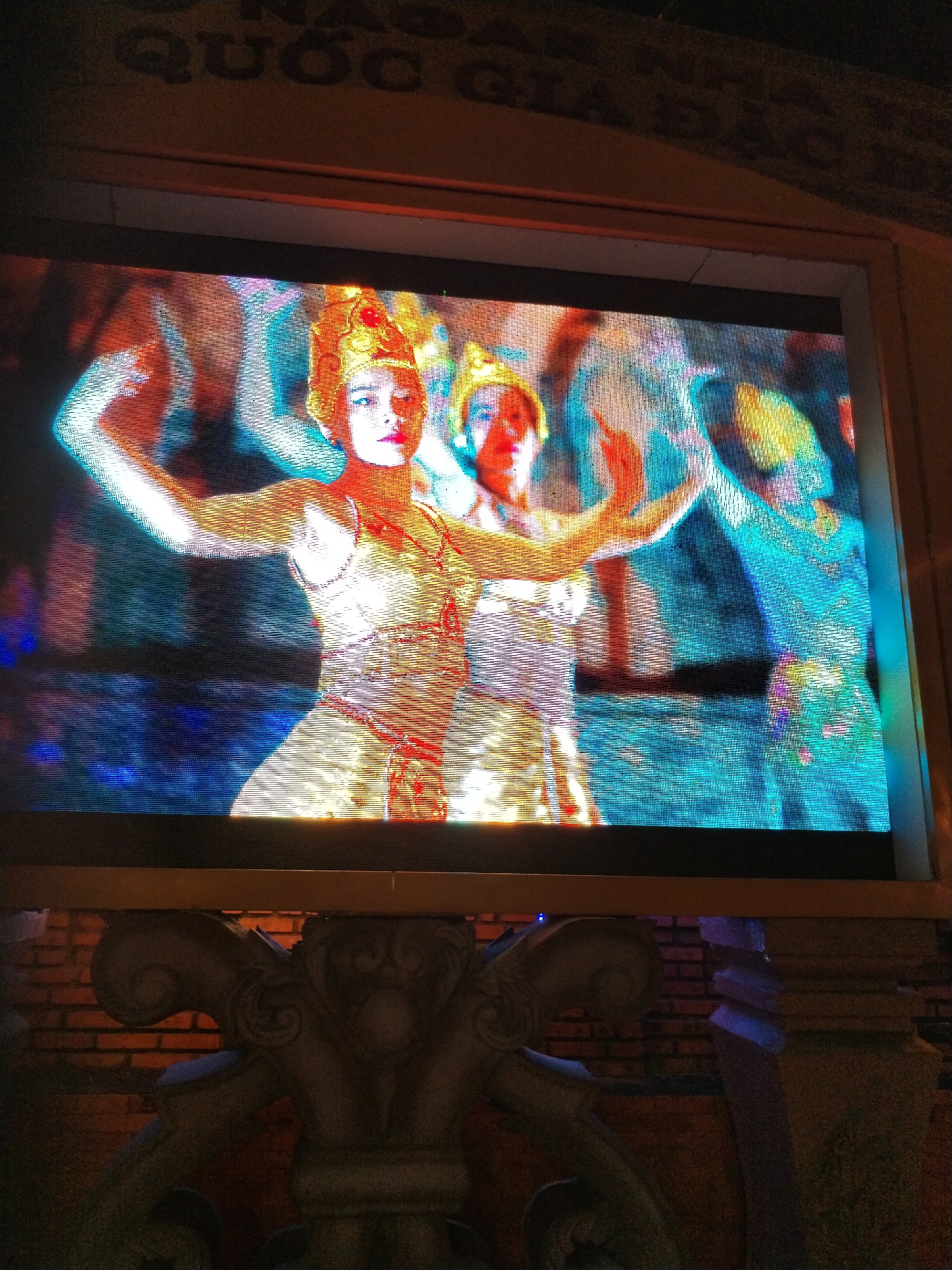 The image shows a television screen displaying a group of dancers in traditional costumes. The dancers wear ornate headdresses and have their arms raised in a dance pose. The screen is mounted on a brick wall with ornate decorative elements below. The setting appears to be in Bac Nha Trang Ward, Vietnam, based on contextual information.