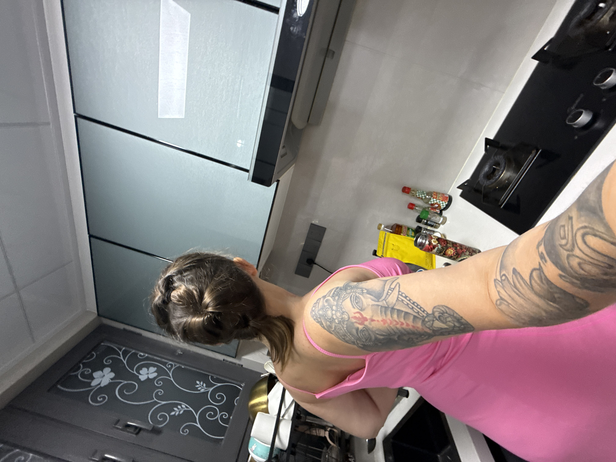 The image depicts a domestic kitchen scene. A person with multiple tattoos on their arms is visible from the back, wearing a pink dress. They appear to be standing and possibly engaged in cooking or preparing food. The kitchen is equipped with a stovetop, cabinetry, and various containers and bottles on the countertop. The overall setting suggests a residential kitchen, potentially in Huizhou, China, given the user's request. The image's composition focuses on the person's back and arms, with the kitchen environment serving as a backdrop.