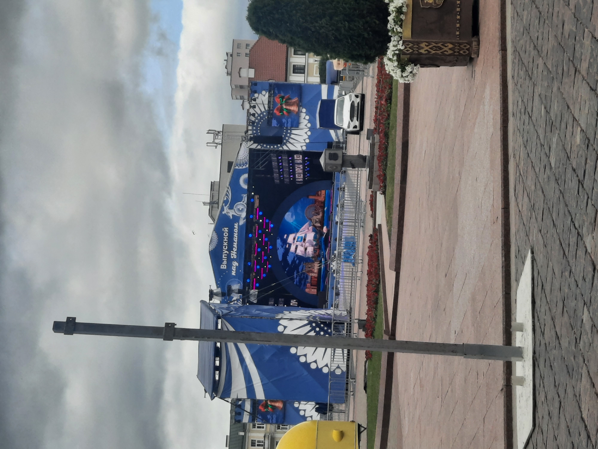 This is an outdoor, eye-level, medium shot of a stage set up for an event. The stage is large and decorated with blue and white banners featuring snowflake-like patterns. The main screen on the stage displays a picture of a sailing ship. There are lights and speakers visible on the stage. A white car is parked near the stage. The event appears to be taking place during the daytime, under a cloudy sky. The ground is paved with light brown and grey bricks. There is a decorative planter with white flowers and a large green tree to the right of the stage. A tall grey pole is in the foreground, partially obscuring the view of the stage. Buildings are visible in the background. Text on the blue banner reads "Выпускной над Неманом," which translates to "Graduation over the Neman." The scene suggests a festive or celebratory event, likely a graduation ceremony, taking place in Hrodna, Belarus, given the reference to the Neman River. There are no people prominently visible in the image.