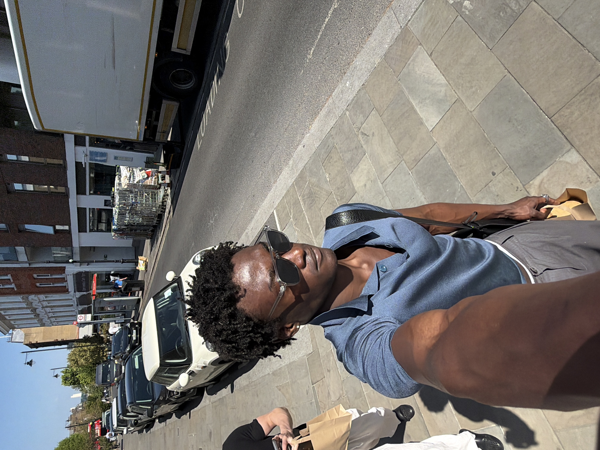 This selfie-style photograph, captured on a sunny day in an urban setting identified as London, United Kingdom, features a man in the foreground. The man, with dark curly hair and dark skin, is wearing dark sunglasses, a light blue collared short-sleeve shirt, and grey shorts. A dark shoulder bag rests on his left side. He is holding the camera to take the picture, with his left hand visible near a light brown paper bag.
Partially visible at the bottom right is another person's lower body and arm, also holding a light brown paper bag, and wearing dark clothing and black shoes.
The background reveals a typical city street scene. A row of parked cars, including a white Fiat 500, lines the left side of the street. Further down, several other vehicles and a large white truck are parked or driving. The street is bordered by multi-story buildings with brick and lighter-colored facades. The ground comprises light grey rectangular pavement slabs on the sidewalk and dark asphalt for the road.
Bright sunlight indicates a clear daytime setting, casting distinct shadows. Visible text on the road reads "LOADING." A red circular traffic sign is also discernible on the left side of the street. The overall impression is a casual moment captured during a stroll in a busy city environment.