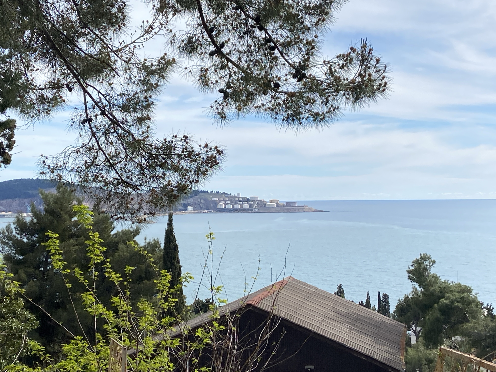 The image shows a coastal view from Šušanj, Montenegro. The foreground includes the partial roof of a building with a dark frame and terracotta tiles. Trees and bushes frame the scene, with a prominent pine branch laden with cones in the upper portion of the frame. The middle ground features the Adriatic Sea, calm and bluish-gray in color. Across the sea, the coastline is visible, and a cluster of industrial structures, likely storage tanks, is situated on the shore. The sky is lightly clouded with a predominantly light blue hue.