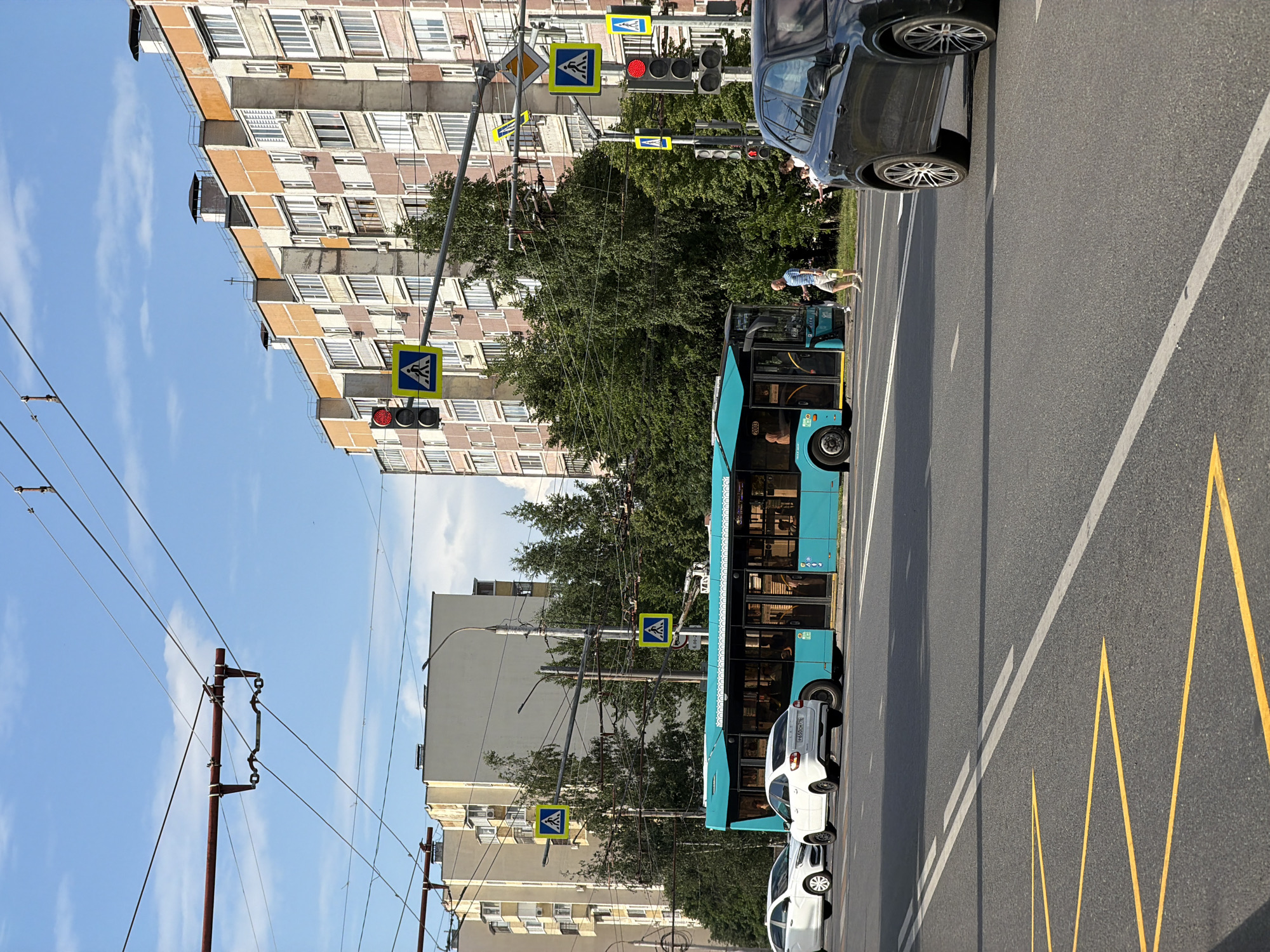 The image shows a street scene in a city under a blue sky with scattered clouds. In the foreground, a black SUV is parked on the right side of the road. The road has yellow zigzag markings and white lane dividers. A bright turquoise bus is stopped at a pedestrian crossing, with several white cars visible behind it. People are walking near the bus and the pedestrian crossing. In the background, tall apartment buildings line the street, with trees interspersed between them. Traffic lights and pedestrian crossing signs are visible, indicating an intersection. Overhead, power lines crisscross the sky.