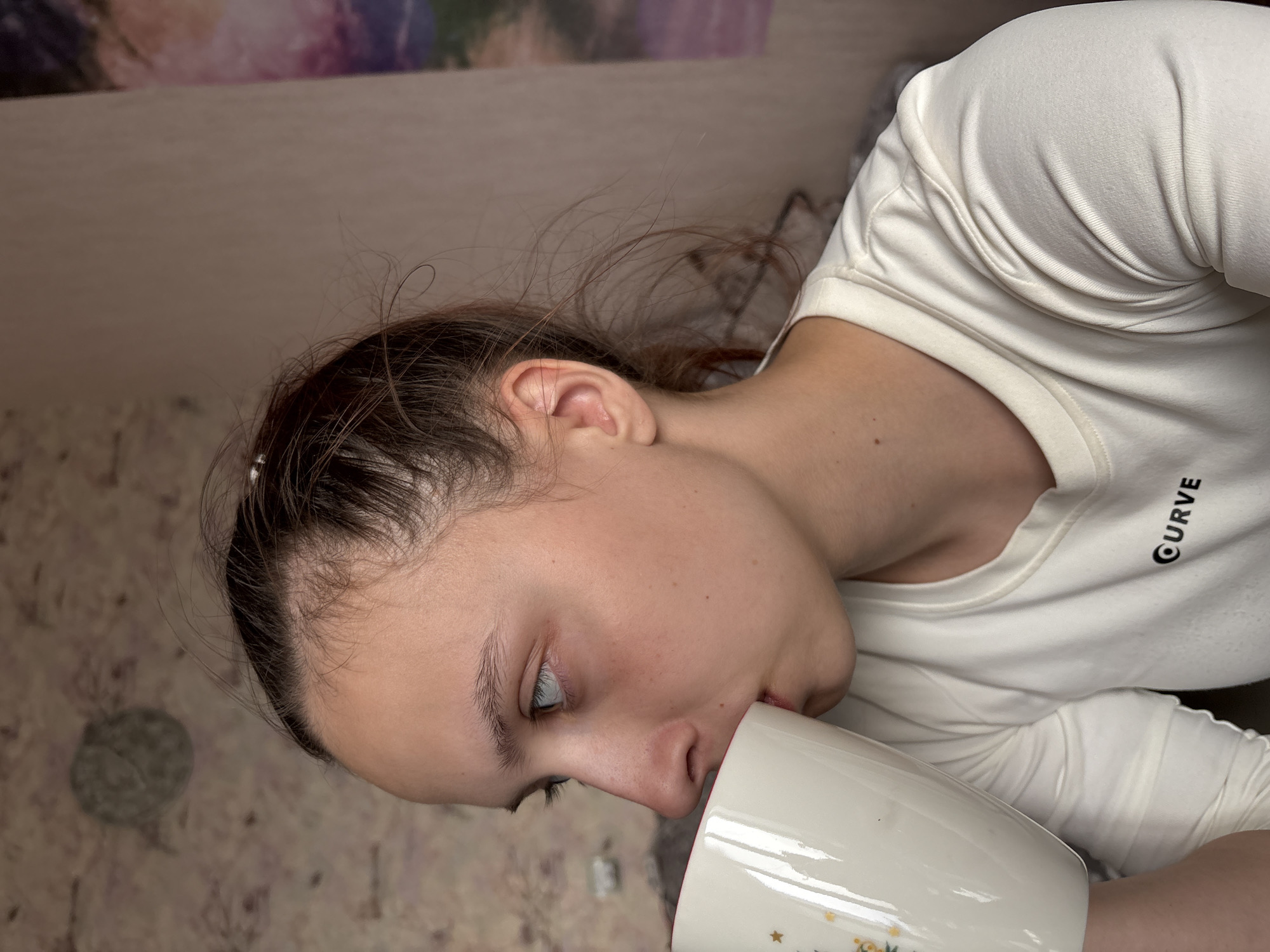 A young person is drinking from a white mug with gold star-shaped decorations. The person has fair skin, brown hair pulled back, and is wearing a white t-shirt with the word "CURVE" printed on it. The background is a blurred wall with a floral pattern and a partially visible image with purple and pink colors. The location is Прикамский, Russia.