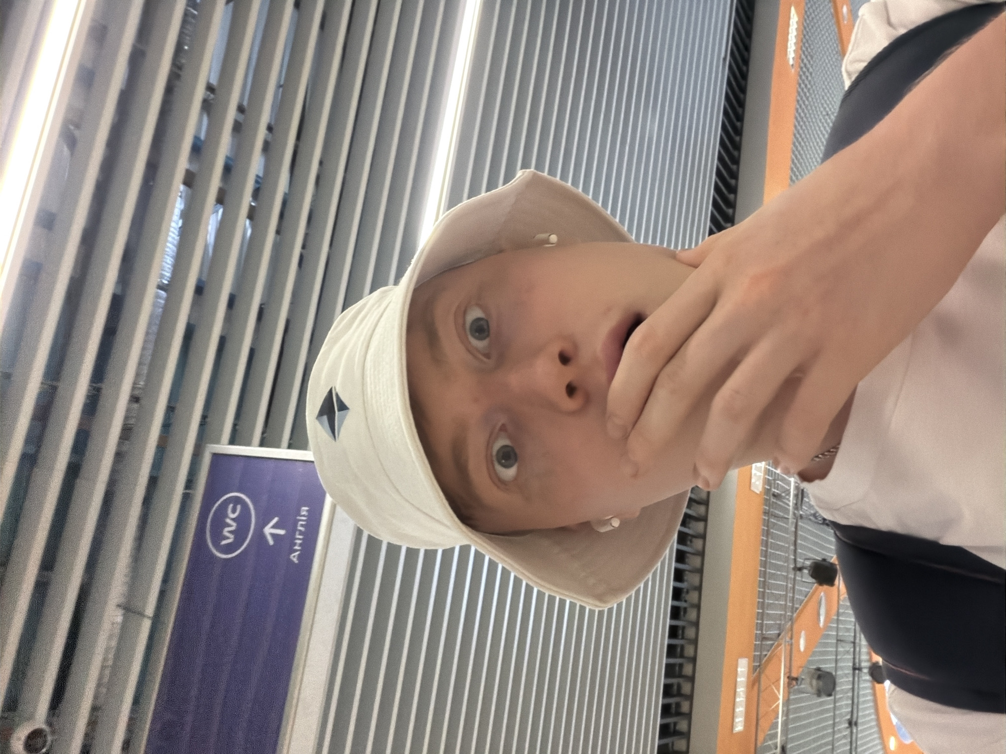 A close-up, selfie-style shot features a young person wearing a white bucket hat and a white t-shirt, with their left hand covering their mouth and their eyes wide open, suggesting surprise or shock. The background includes a sign with "WC" and an arrow pointing upwards, indicating restrooms, with Cyrillic text below it that reads "Англiя" (Anglia, meaning England). The environment appears to be an indoor public space with a metallic, slatted ceiling and orange accents visible in the background, possibly an airport or a large public building. The lighting suggests artificial indoor illumination.