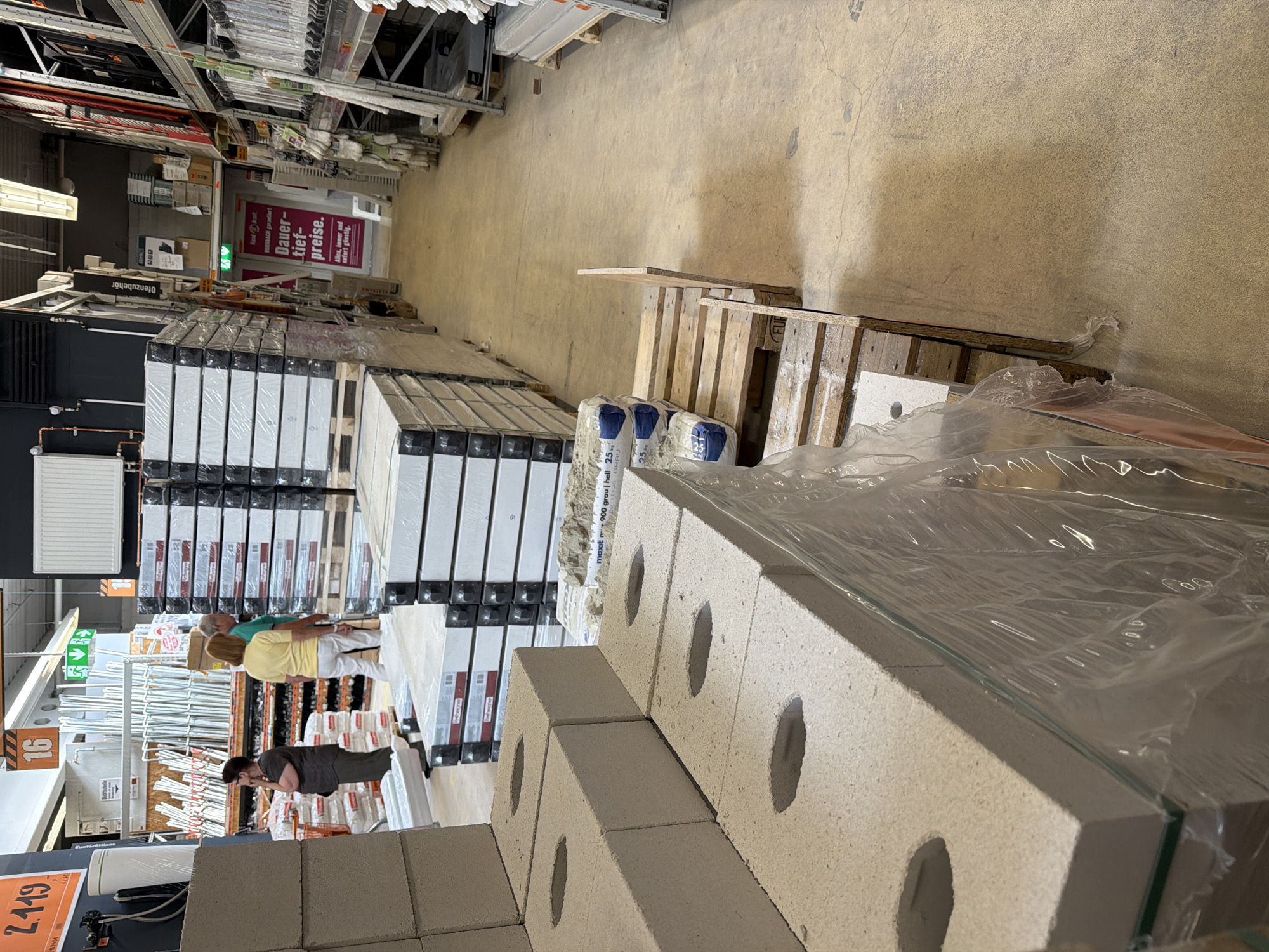 This image appears to be taken inside a hardware store, likely in Gütersloh, Germany, given the German language signs. The store is well-stocked, with shelves filled with various construction and hardware items, including what seems to be piping, insulation materials, and stacks of building blocks wrapped in plastic. At the foreground, there's a prominently displayed stack of concrete blocks with circular holes.
Several people are present in the middle ground, browsing the merchandise. There are two women wearing yellow and green tops respectively, and one person with a gray T-shirt, inspecting the products.
Signs in the store advertise "Dauer-Tief-Preise" (long-term low prices), indicating special offers. The image's composition, with the stacks of materials and people moving through the aisles, suggests a typical retail environment for construction and home improvement goods.