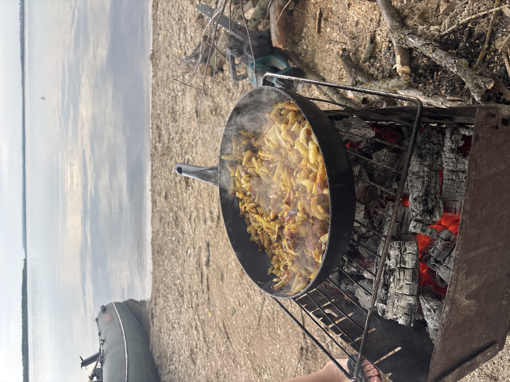 The image depicts an outdoor cooking scene on a sandy or dirt shoreline, identified as Unknown, Unknown. In the foreground, a black frying pan containing a quantity of fried potatoes and onions, with visible steam emanating, is positioned on a metal grill rack. This grill rack sits atop a portable, rectangular metal brazier filled with glowing red coals and grey ash. To the immediate left of the brazier, the left foot of an individual, wearing a dark sandal, is partially visible.
The background features a wide, calm body of water, reflecting the overcast sky with light clouds. On the left side of the water, an olive-green inflatable boat with a dark upper tube is partially beached. To the right of the cooking setup, a dark-colored chainsaw with a light blue housing is visible lying on the ground amidst an accumulation of wood shavings, cut branches, and logs, suggesting recent wood-cutting activity. The overall lighting indicates an overcast day.