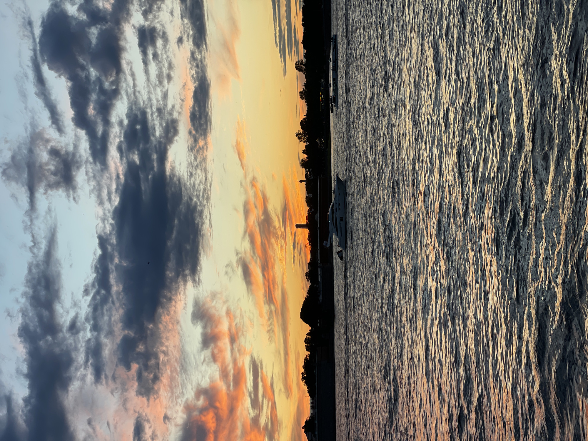 This media file is a landscape photograph captured at sunset. The image is divided into two distinct halves: the left side depicts a dramatic sky filled with dark, swirling clouds tinged with vibrant orange and yellow hues from the setting sun. The right side shows a body of water, likely a lake or ocean, with its surface rippling and reflecting the golden light of the sunset, creating a textured, shimmering effect.
Along the horizon, a silhouetted tree line separates the sky from the water, suggesting a coastal or lakeside setting. A few boats are visible on the water, one larger yacht in the foreground and others further in the distance. A tall, slender structure, possibly a lighthouse or tower, is also faintly visible on the distant shore. The overall mood of the image is serene and tranquil, amplified by the warm, fading light and the gentle movement of the water. The time of day is clearly sunset, indicated by the colors in the sky and the lighting on the water. There is no visible text in the image.