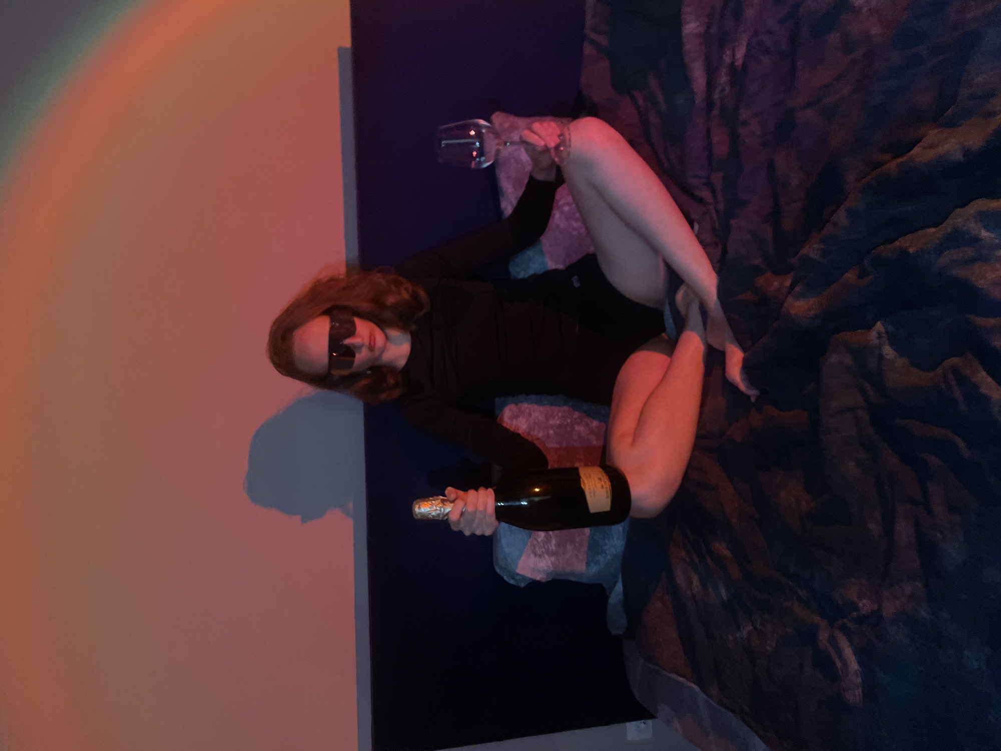 The image depicts a woman lying on a bed. She is wearing a dark outfit and sunglasses. She is holding a champagne bottle in one hand and a wine glass in the other. The room has a colorful, possibly artificial, lighting effect. The scene is indoors, possibly in a private residence. There is no information that can be used to place this at the location of the city of Подшивалово, Russia.