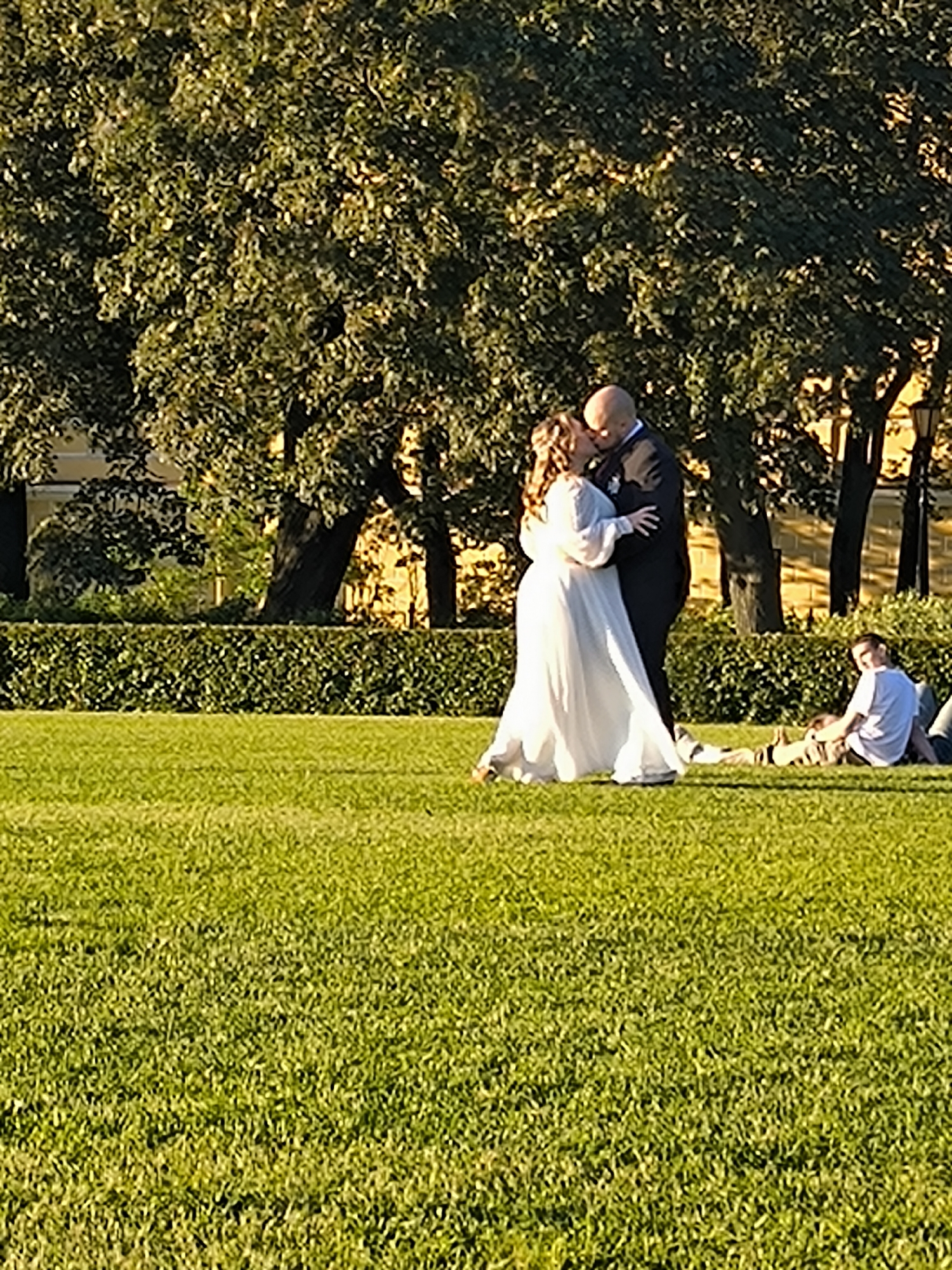 In a park-like setting, a newlywed couple is captured in a tender moment, sharing a kiss. The bride, adorned in a flowing white gown, stands with her arms around the groom, who is dressed in a dark suit. Their romantic interlude unfolds on a lush green lawn, bordered by a manicured hedge and mature trees. In the background, a hint of a building with warm-toned walls suggests the presence of urban architecture. To the right, two individuals are relaxing on the grass, one sitting up and observing the scene. The afternoon sun casts long shadows, bathing the park in a golden light.
