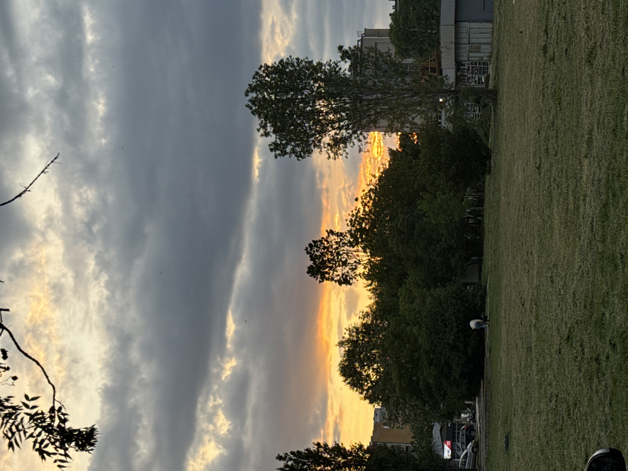 The image shows a park in Sofia, Bulgaria at dusk. The sky is filled with grey and white clouds, and the setting sun casts a golden glow on the horizon. Trees line the park, with their silhouettes visible against the light. A grassy field stretches into the distance, where cars are parked along a street. A building is visible in the background. An individual bends over near the edge of the grass.