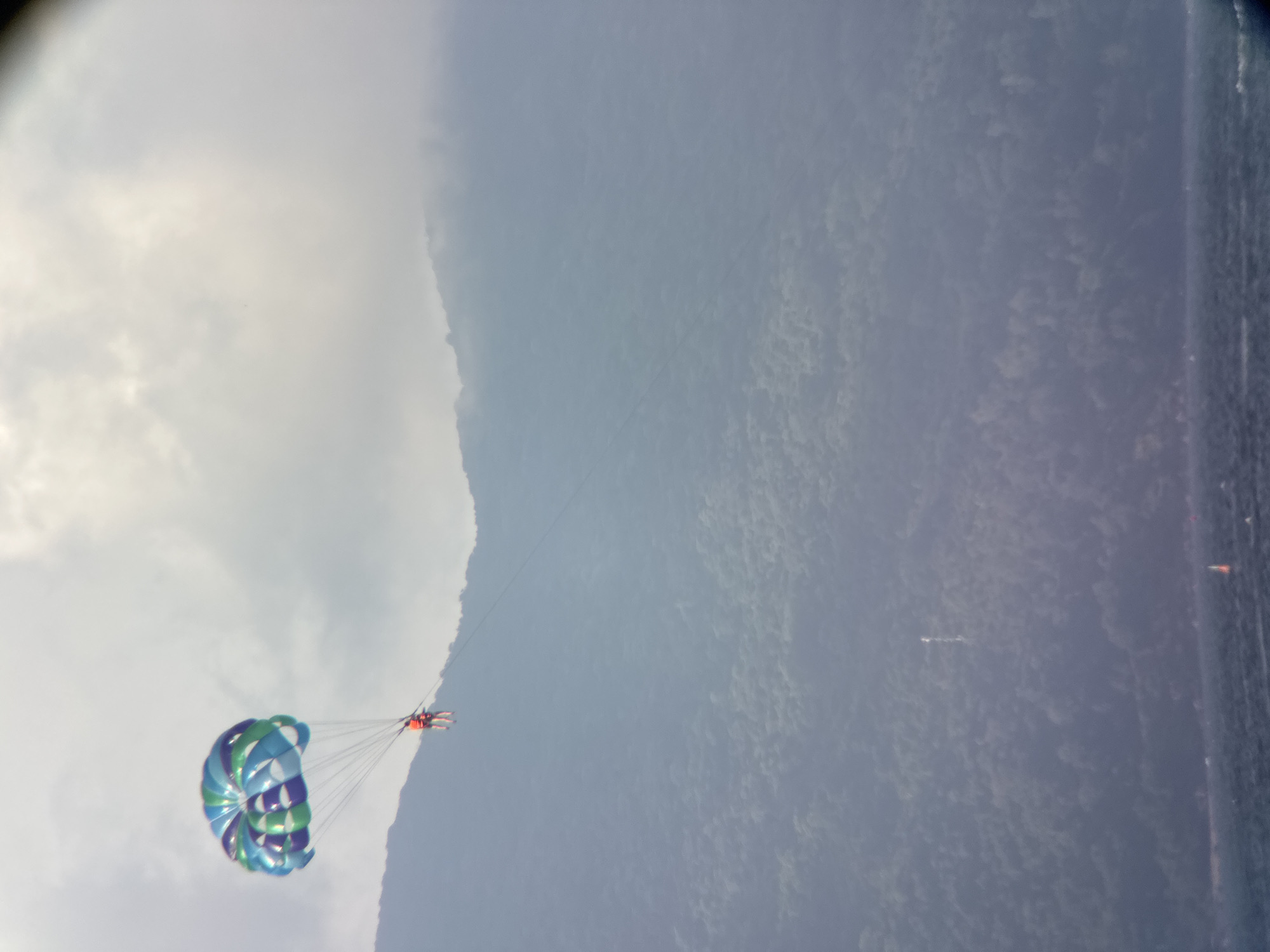 Two individuals are parasailing over the ocean with a lush, forested mountainside in the background. The parasail is colorful, with sections of blue, green, and purple. The individuals are wearing orange life vests and are suspended in the air, attached to the parasail by ropes. The mountainside appears to be covered in dense vegetation and is partially obscured by clouds or mist. The water in the foreground is choppy, and a small orange buoy is visible in the distance. A distant tower-like structure is visible on the mountainside. This scene is typical of tourist activities offered along the coast of Đà Nẵng, Vietnam, which is known for its scenic beaches and mountainous backdrop.