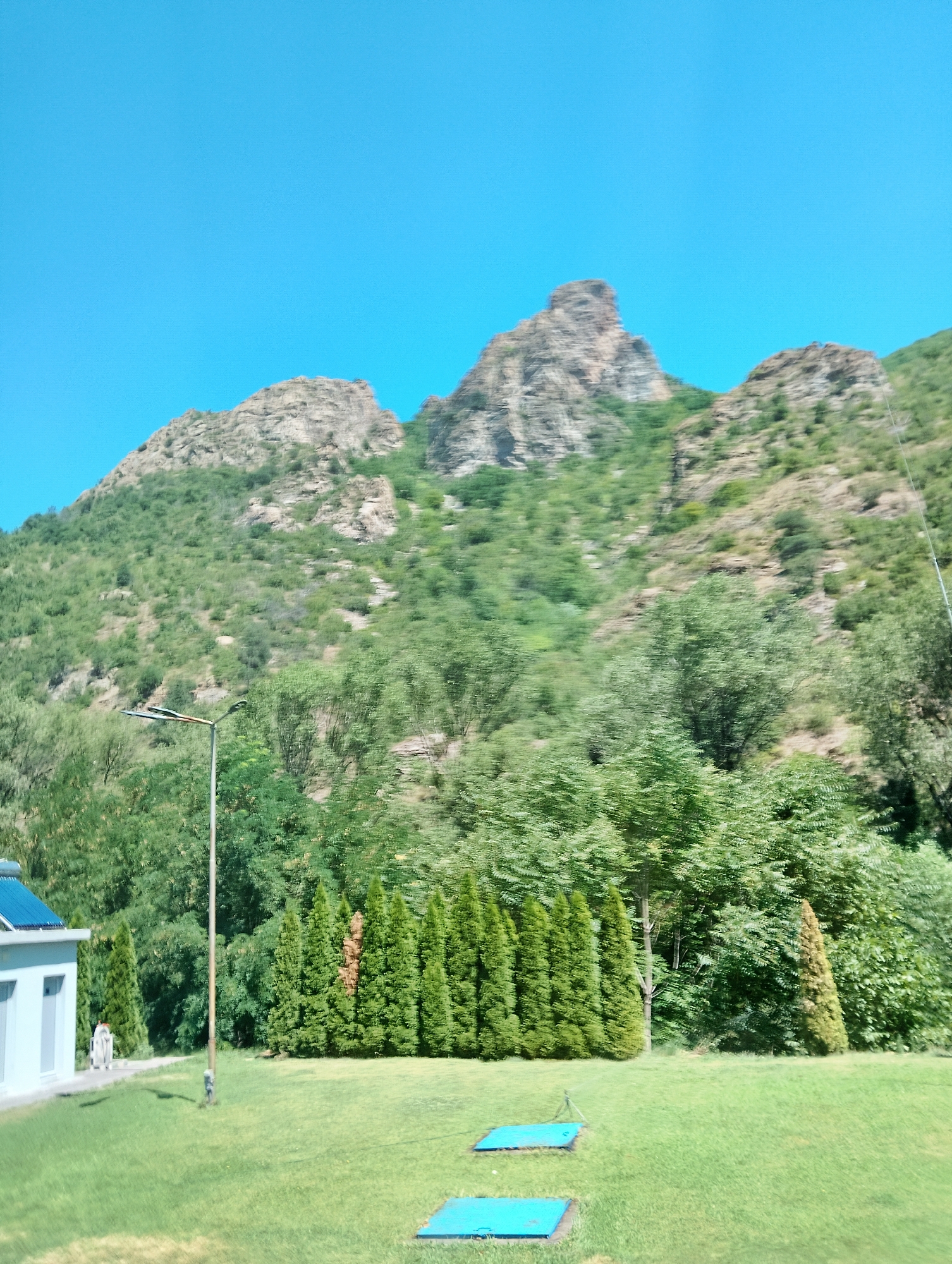 The image depicts a natural landscape dominated by a large, rocky mountain covered in lush green vegetation. The mountain has rugged peaks and slopes, with dense trees and shrubs growing on its lower sections. In the foreground, there is a well-maintained grassy lawn. A light pole with a modern fixture stands on the left side of the lawn. Adjacent to the lawn, on the far left, is a light-colored building with a blue roof and white window frames. Two rectangular blue structures are visible on the grass in the middle ground. A line of tall, conical evergreen trees forms a partial border on the right side of the lawn. The sky above is a clear, bright blue. The image appears to be taken from a slightly elevated perspective, and there is a sense of motion blur in the foliage, suggesting the photo was taken from a moving vehicle. This scene is located in Krupnik, Bulgaria.