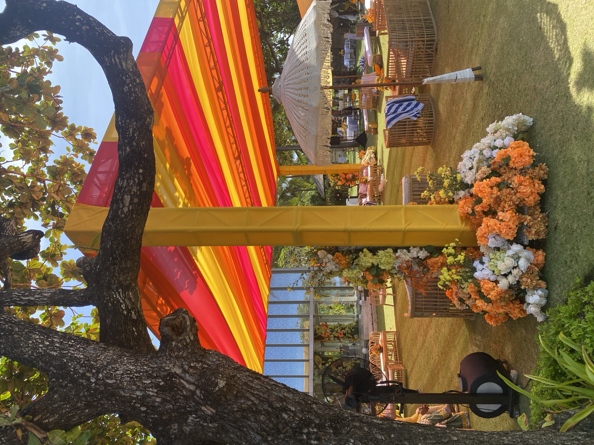 The image shows an outdoor event setup in Kuta Selatan, Indonesia. The scene includes a colorful canopy with stripes of pink, orange, and yellow, supported by yellow poles. Under the canopy, there are arrangements of wicker furniture, white decorative umbrellas, and floral displays with orange and white flowers. In the background, there appears to be a structure with glass panels. A large tree with thick branches and foliage is prominently positioned in the foreground, partially obscuring the view. An industrial fan and lighting equipment are also visible near the base of the tree. The setting is on a green lawn, suggesting a garden or park environment.