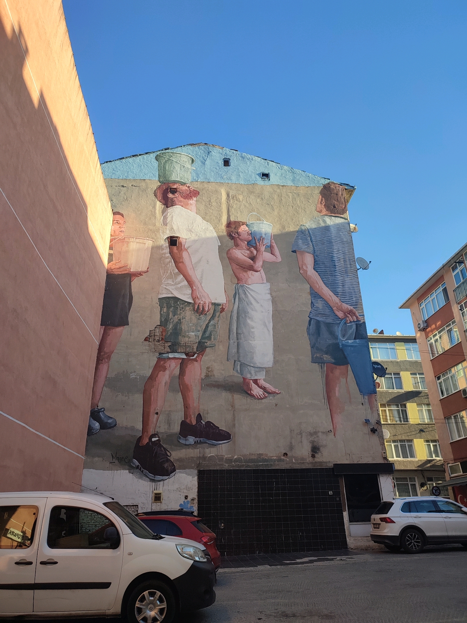 The image shows a mural painted on the side of a building in Kadıköy, Turkey. The mural depicts several figures carrying buckets. The building itself is painted in shades of beige and blue, and it is situated in a narrow street with other buildings visible in the background. A white van and a red car are parked in front of the building. The sky is clear and blue. The mural appears to be located on Yeldeğirmeni Sokak in Kadıköy, a neighborhood known for its street art.