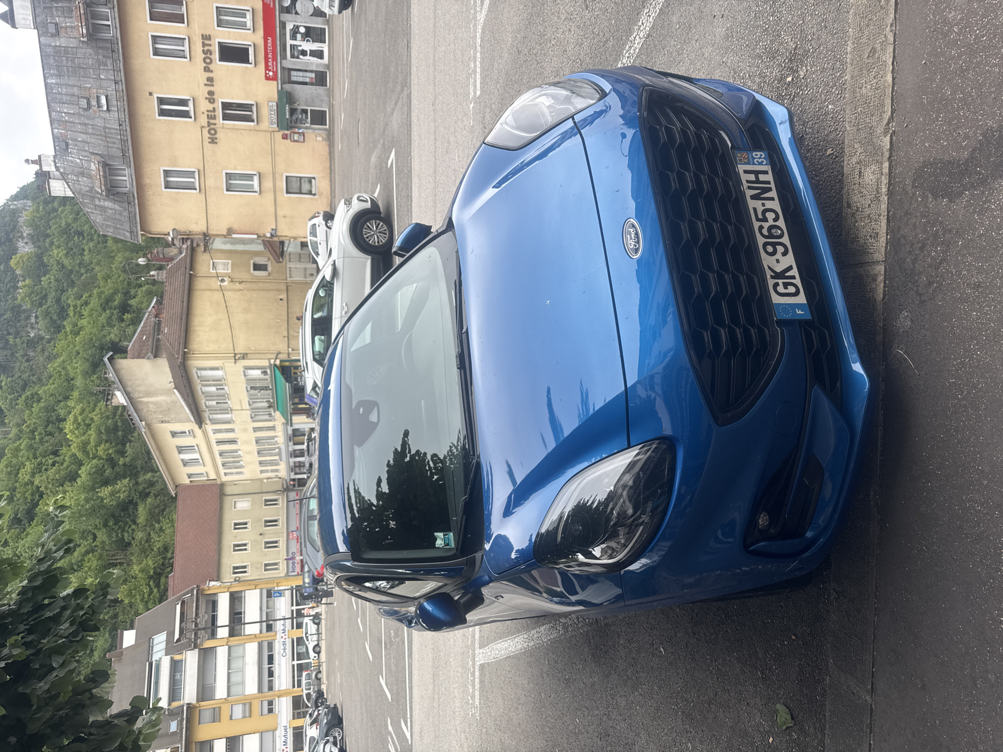 A blue Ford Puma is parked in a paved lot in front of buildings in Saint-Claude, France. The car has a license plate that reads "GK-965-NH 39". In the background, there are several buildings, including one with "HOTEL de la POSTE" written on it, and a steep, wooded hillside. The sky is overcast.