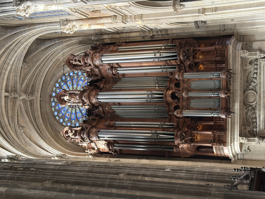The image displays the grand pipe organ situated within the Saint-Eustache Church in Paris, France. The massive instrument dominates the central frame, constructed from dark, intricately carved wooden casing and numerous metallic pipes varying in length and diameter, arranged in symmetrical vertical clusters. The organ features multiple tiers of ornate architectural detailing, including columns, pilasters, balustrades, and elaborate carvings. Several sculpted figures, appearing to be statues of individuals, are positioned on prominent pedestals along the upper sections of the wooden casing. Directly above the organ, a large circular rose window with stained glass radiates blues, reds, and yellows, providing ambient light. The surrounding environment consists of the church's interior, characterized by tall, fluted stone columns, ribbed vaulted ceilings, and pointed arches, all constructed from light-toned stone. Natural light illuminates the scene, highlighting the architectural details. No people are visible, and no actions or interactions are depicted.