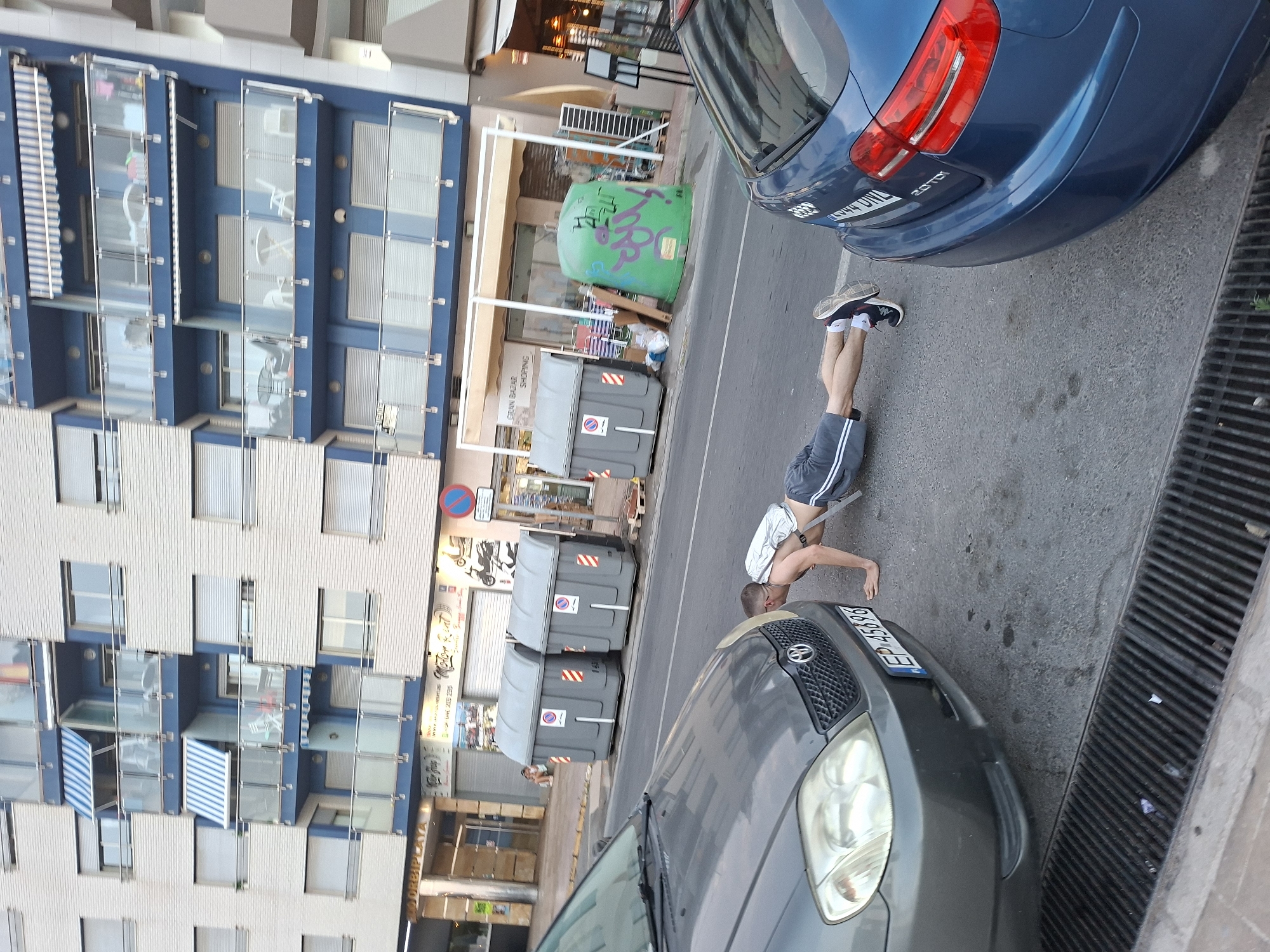 The image depicts a street scene in Torrevieja, Spain. In the foreground, a person is performing a push-up on the asphalt. They are wearing grey shorts and a white shirt. The person's legs are bent at the knees, and their feet are slightly elevated, suggesting a modified push-up or perhaps part of a different exercise. Behind the person, a gray car is parked, partially obscuring the view. Further back, two large gray garbage bins are visible, one with a red and white striped sticker. Next to the bins, a large, bright green cylindrical object, possibly a larger waste container or a decorative element, is covered in graffiti.
The background features a multi-story building with a light-colored facade, featuring numerous windows and blue accents. Awnings are visible over some of the shops at the ground level. The overall scene suggests an urban environment, likely a commercial or residential street.
The lighting indicates it is daytime, with shadows cast on the ground. The weather appears to be clear, as there are no visible signs of rain. The presence of graffiti and the general street clutter suggest a lived-in, possibly bustling, urban area. The license plate on the gray car reads "EP 5586". Another car, a blue one, is visible in the upper right portion of the image. The image is taken from a slightly elevated, angled perspective, looking down towards the street.