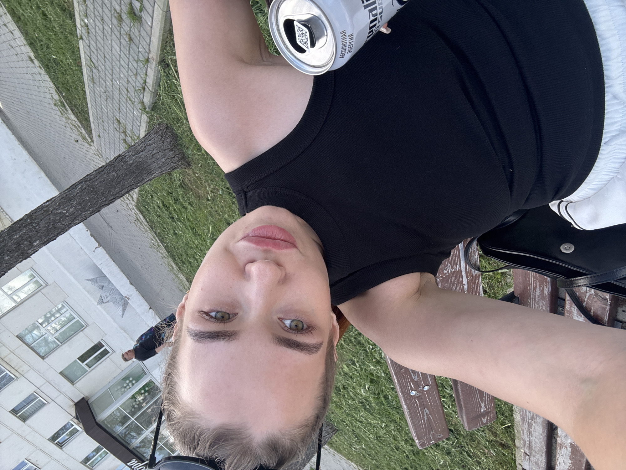 A woman is centered, holding a can, wearing a black shirt and white pants, seated on a bench outdoors. A building is visible in the background. A person is visible behind her and in the background near the building. The location is the city of Chaikovsky, Russia.