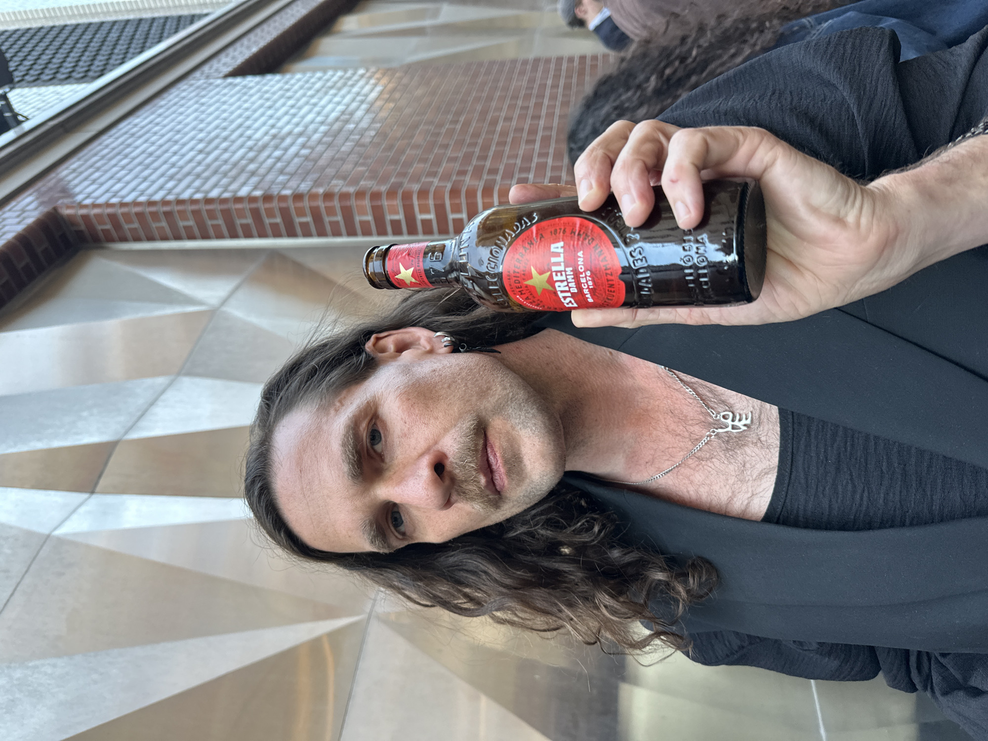 An upright portrait-oriented image shows a man holding a bottle of beer, looking towards the viewer in an urban setting. The man has long, dark, wavy hair, a mustache, and light-colored eyes, with a neutral to contemplative expression. He wears a dark shirt and jacket, and a silver chain with a unique pendant around his neck. An earring is visible in his left ear.
He holds a dark glass bottle with a prominent red and gold label in his right hand. The label clearly reads "ESTRELLA DAMM" in gold lettering, with "BARCELONA" and "1876" also visible, indicating the brand is Estrella Damm, originating from Barcelona. Other partially visible text includes "ORIGINAL" and "MEDITERRANEAN".
The background features contemporary architecture, with a section of a brick wall visible on the right, and a sleek, light-colored geometrically patterned wall behind the man and to his left. The lighting suggests it is daytime. A blurry figure is barely discernible in the distant background. The location is an urban environment in London, United Kingdom.