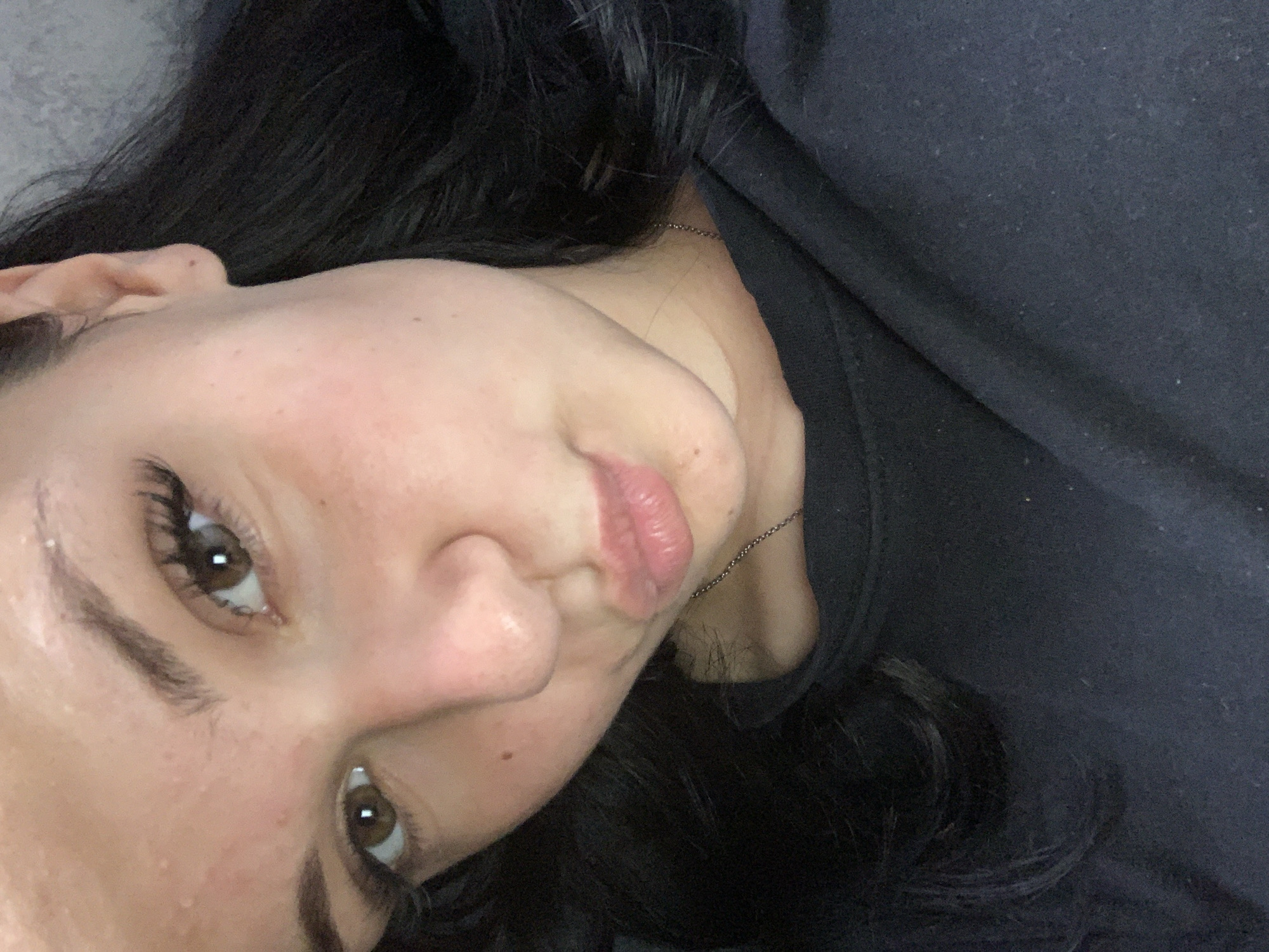 A close-up selfie shows a young woman with dark hair and brown eyes. She is wearing a black top and a delicate silver necklace. Her face is tilted slightly, and her lips are pursed in a kiss. The background is out of focus, but appears to be an indoor setting. The image was taken in the city of Ustyvytsia, Ukraine.