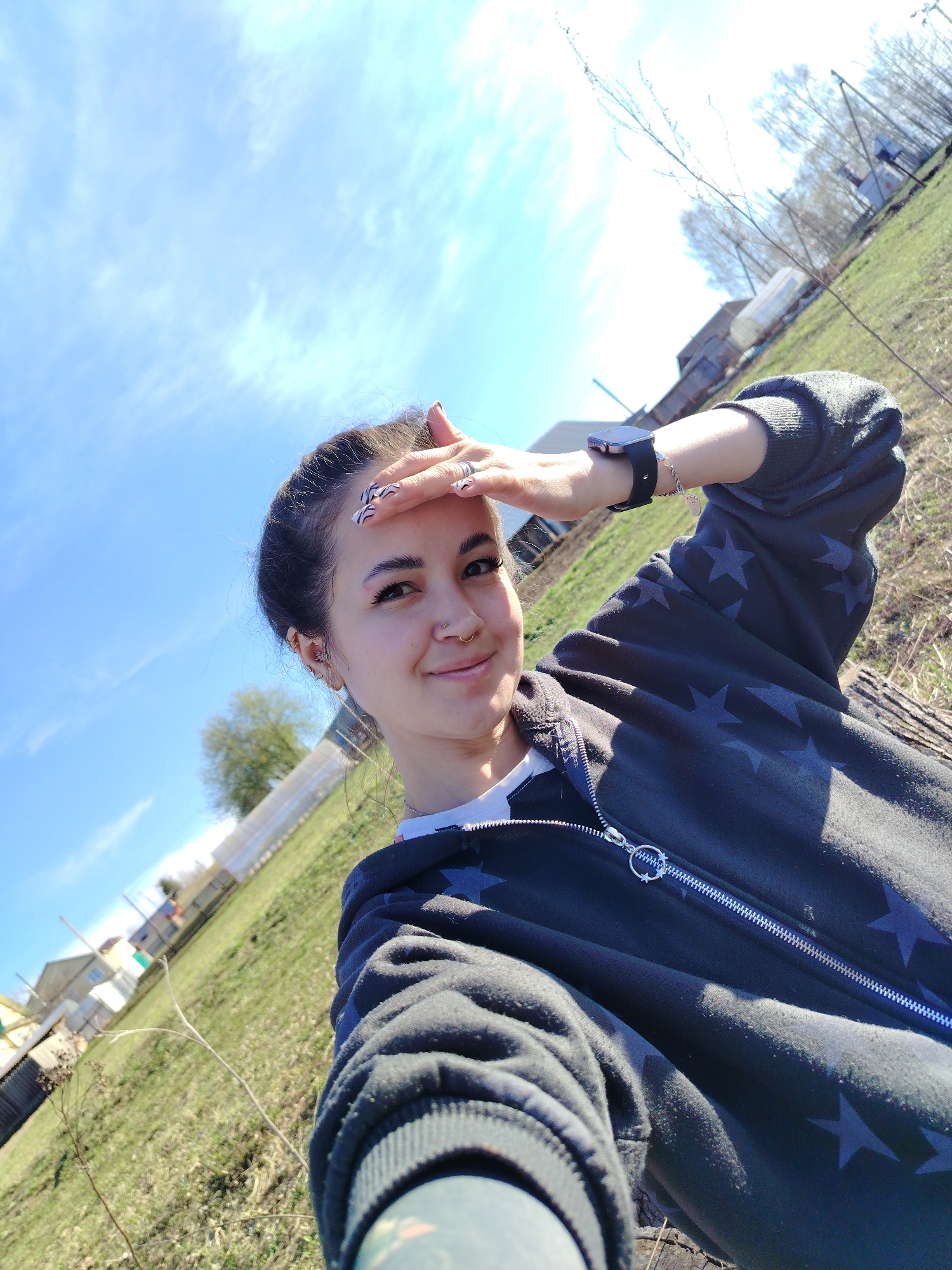 A young woman is taking a selfie outdoors in Гожан, Russia on a bright, sunny day. She is fair-skinned with dark hair pulled back and adorned with patterned nails. She wears a dark-colored zip-up hoodie patterned with stars, layered over a white top. The woman has multiple piercings on her ears, a nose ring, and is wearing a smartwatch on her left wrist. She is shielding her eyes with her hand, looking into the camera with a slight smile. The background shows a rural scene with a slightly tilted horizon, featuring a grassy area, scattered trees, and houses in the distance. The sky is blue with scattered white clouds.