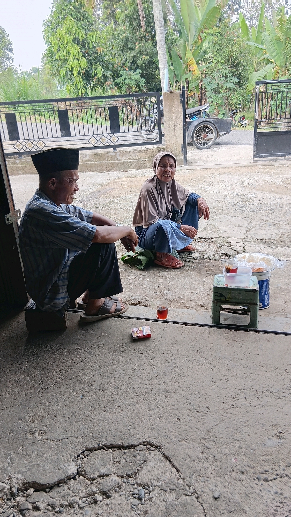 Two individuals are sitting on the ground in an outdoor setting. A man, wearing a dark cap and a striped shirt, is seated on a dark rectangular object. Beside him, a woman in a headscarf and blue pants is crouched on a green leaf. In the foreground, a pack of cigarettes and a glass of amber liquid are placed on the concrete ground. Behind them, a black metal gate with decorative elements and a motorcycle with a trailer are visible. The background features lush green trees and vegetation.