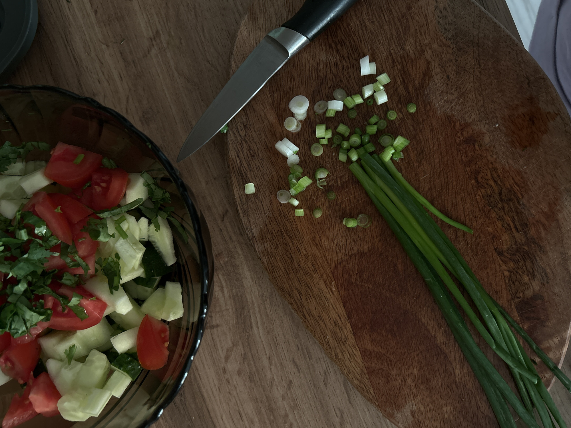 A high-angle medium shot shows a close-up of chopped green onions on a wooden cutting board next to a knife and a bowl of salad. The salad bowl, made of dark brown glass, contains chopped tomatoes, cucumbers, and green herbs. The cutting board is made of reddish-brown wood with visible grain and scratches. A silver chef's knife with a black handle lies on the board, pointing towards the salad. Chopped green onions are scattered across the board, with a bunch of uncut green onions lying to the lower left. The background is a wooden table surface. The lighting suggests an indoor setting.