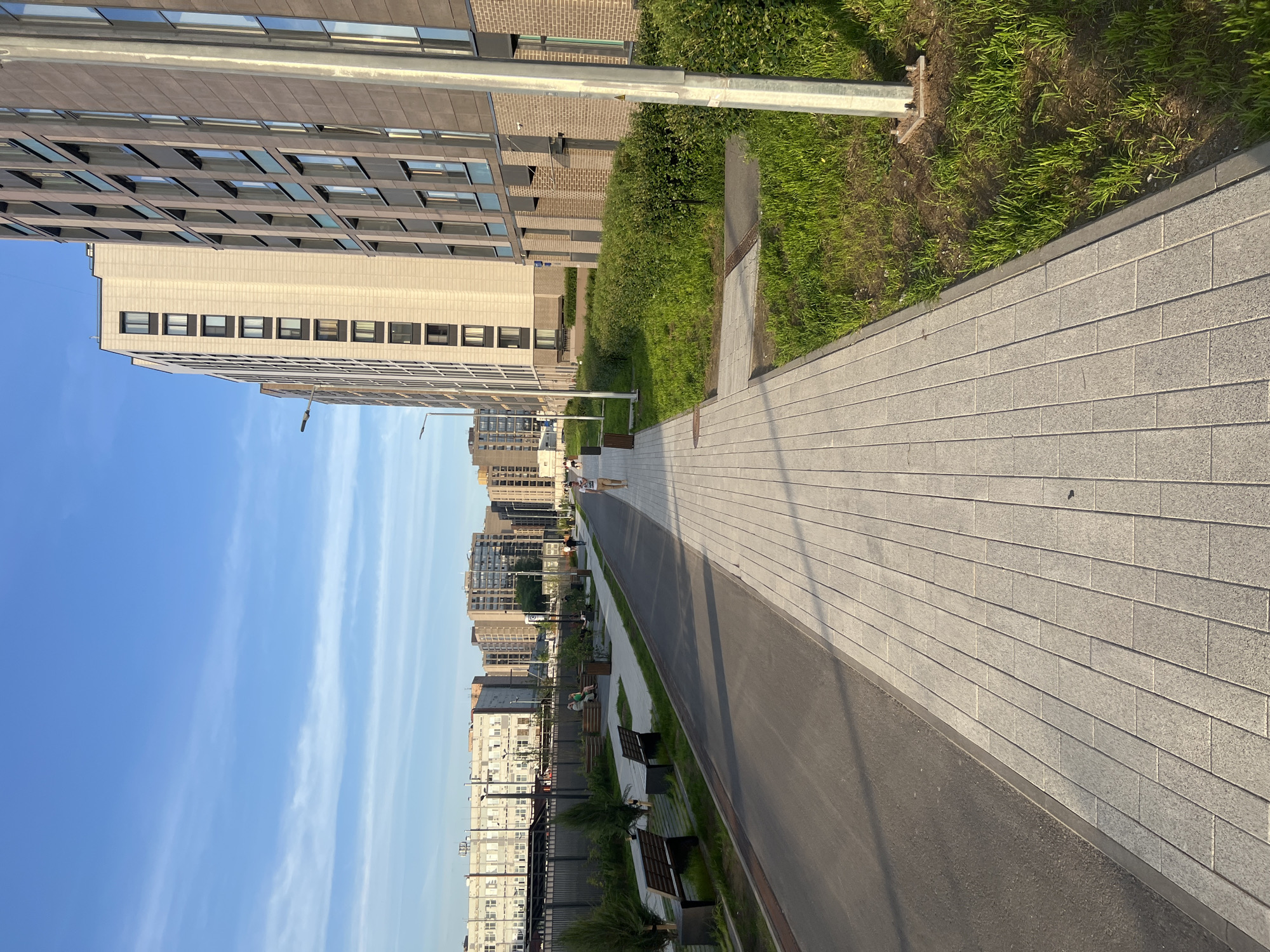 This image depicts a modern urban scene in Saint Petersburg, Russia, showcasing a pedestrian and bicycle thoroughfare flanked by contemporary architecture and landscaped green spaces, illuminated under a clear blue sky with sparse, wispy clouds.
On the right, a row of tall, multi-story residential buildings dominates the frame. The closest building features a dark brown/grey facade with numerous rectangular windows and vertical grey structural elements. Adjacent to it, a lighter, cream-colored building stands, characterized by an array of dark-framed windows. A low brick wall base and landscaped bushes separate these buildings from a wide, light grey paved sidewalk, constructed from rectangular pavers laid in a staggered pattern. A single, tall grey lamppost is prominently visible in the far right foreground.
To the left of the paved sidewalk, an asphalt path, likely for bicycles or service vehicles, runs parallel. This path is bordered on its left by a narrow strip of grass and small ornamental trees or shrubs planted within square cutouts. Beyond this vegetation strip, a dark metal fence separates the pathway from an older, multi-story industrial or commercial building with numerous white-framed windows, contrasting with the modern residential structures. Further in the background on the left, additional modern residential buildings are visible.
Several individuals populate the scene. One person, dressed in a light-colored top and bottoms, walks away from the camera along the main paved sidewalk in the mid-ground. On the left side of the asphalt path, two people are seated on a dark wooden bench, appearing to interact, with one wearing a green top. Another wooden bench is visible closer to the foreground on the left, and additional benches and dark trash receptacles are placed intermittently along the path. Streetlights are positioned at regular intervals along both sides of the pathways.
The natural light suggests either early morning or late afternoon, with the sun casting long shadows, indicating clear weather. The ground areas consist of neatly maintained grass patches and low, dense green shrubbery.