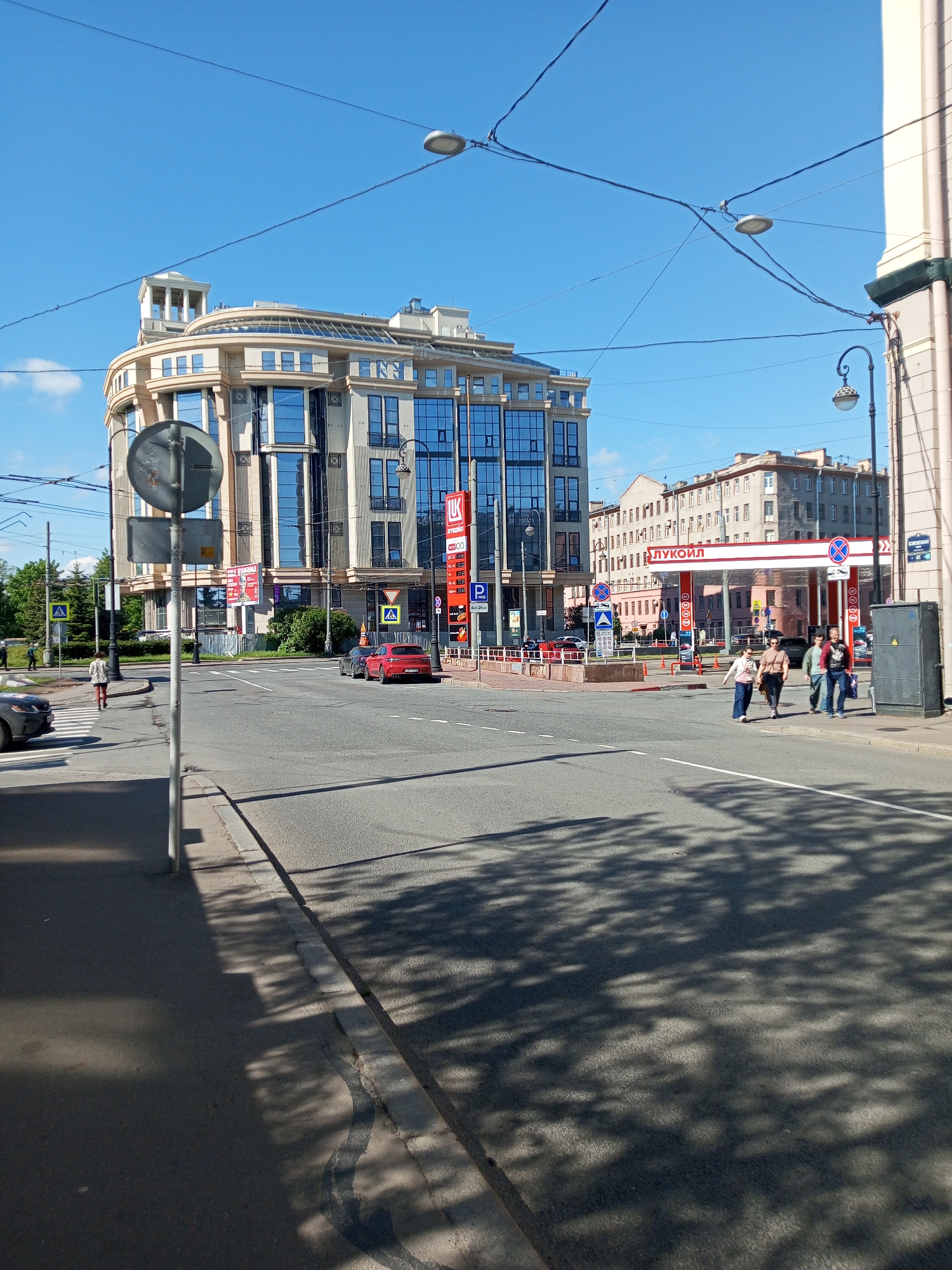 The image shows a street scene in Saint Petersburg, Russia, on a sunny day. The most prominent feature is a modern office building with a curved façade and glass windows. Across the intersection is a Lukoil gas station. Several people are walking across the street. Cars are parked on the side of the road. Traffic signs and overhead wires are visible. The shadow of trees is cast on the road.