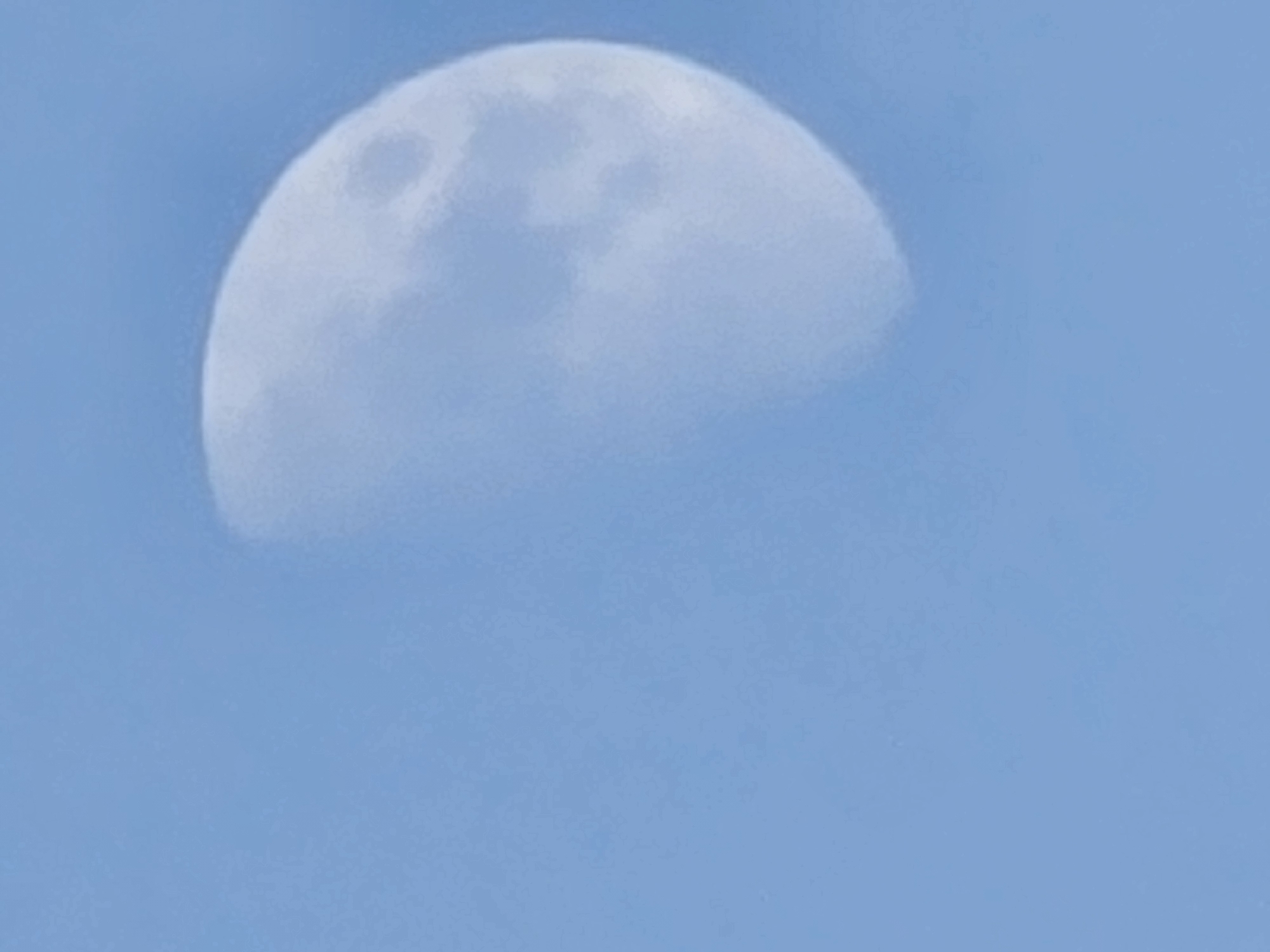 A half-illuminated moon is visible against a clear blue sky. The moon appears in the upper right quadrant of the image, with its bright, circular portion illuminated and its shadowed portion curving towards the left. Subtle variations in the moon's surface suggest craters and maria. The sky is a uniform, soft blue, devoid of clouds or other celestial bodies. The image appears to have been taken from an outdoor location within the city of Podgorica, Montenegro, at an unspecified time of day.