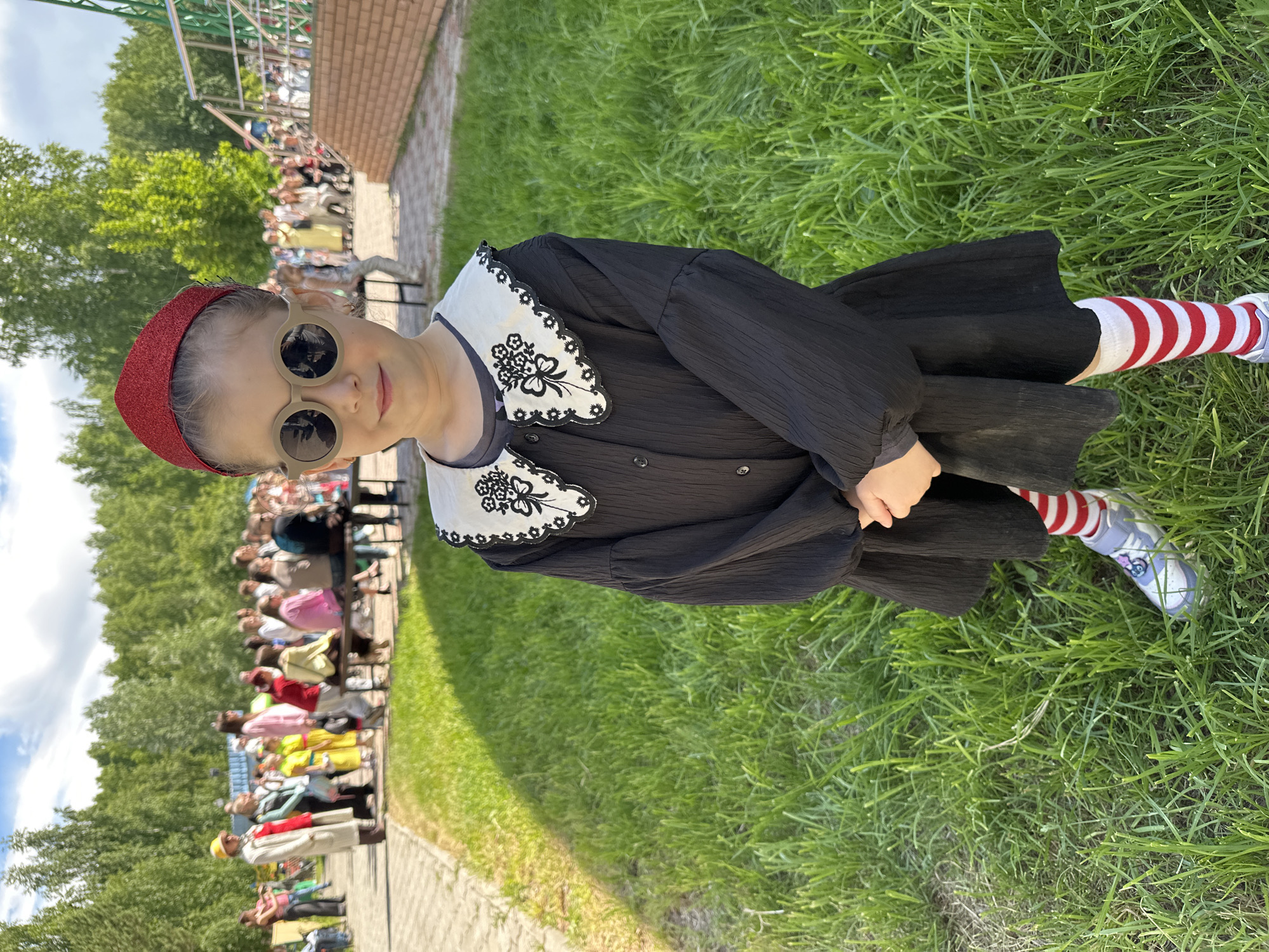 A young girl stands on a patch of green grass near a paved walkway. She is wearing a black dress with a white lace collar, a red headpiece, round sunglasses, and red and white striped socks with light blue shoes. In the background, a group of people sits on benches, watching something. Trees and a brick building are also visible. The location is in the city of Chaikovsky, Russia.