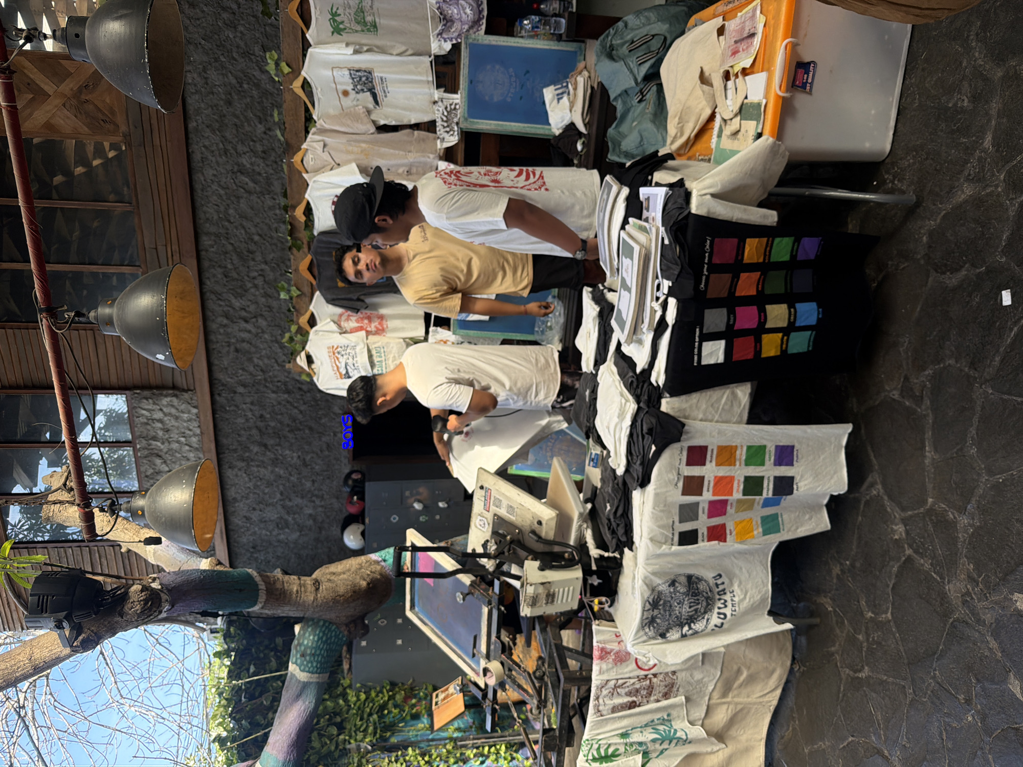 The image shows an outdoor screen printing stall in Kuta Selatan, Indonesia. Two people are standing behind a table covered with stacks of T-shirts. The table displays designs and color swatches. A screen printing setup is visible, including screens and equipment. T-shirts are hanging on racks in the background. The scene is situated outdoors with trees and a building in the background.