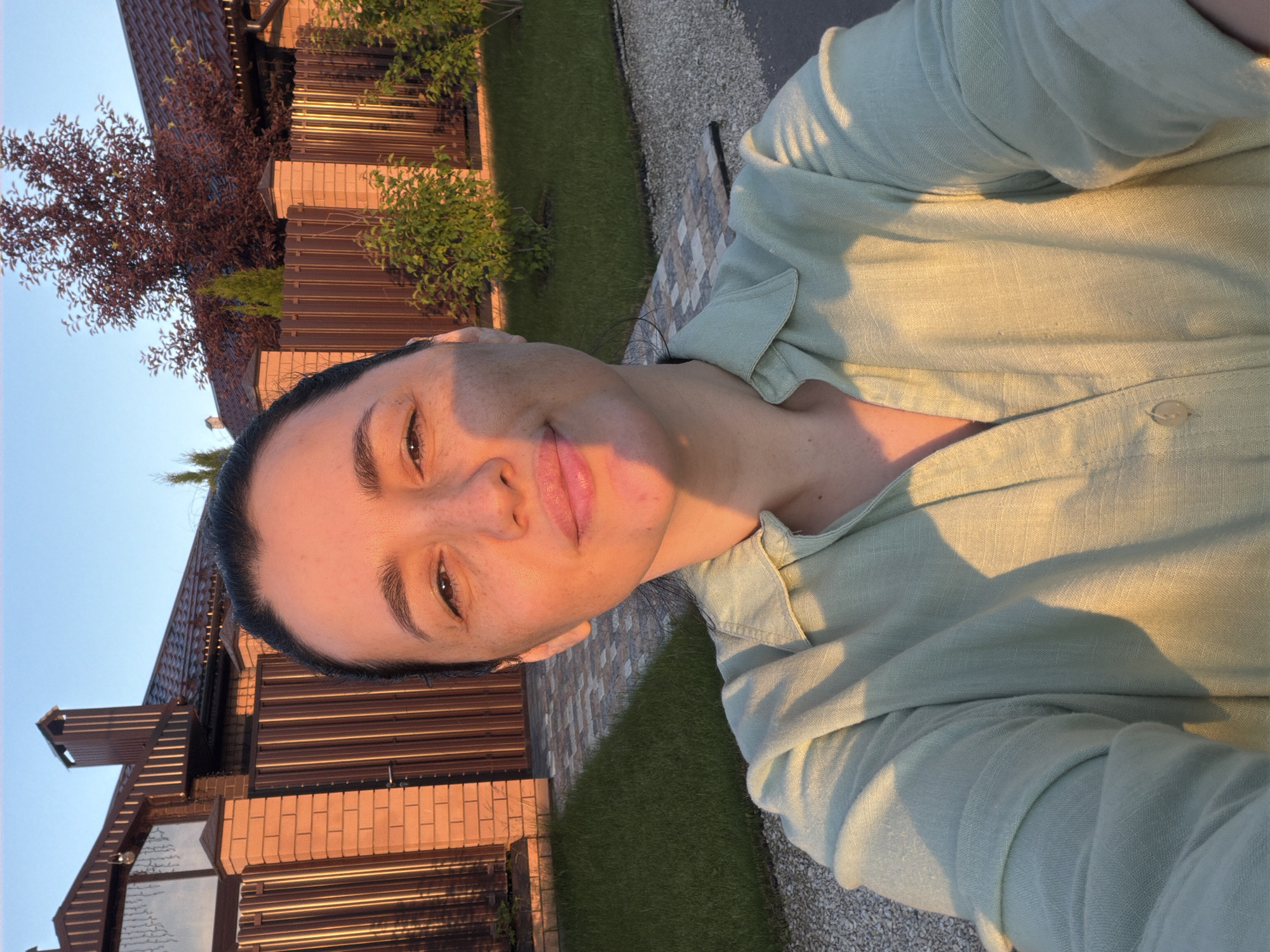 A woman with dark hair and a light green collared shirt takes a selfie outdoors. She is positioned in the foreground, with a brick and metal fence, a house with a brown roof, and a tree visible in the background. The setting appears to be a residential area. The lighting suggests it is either sunrise or sunset, casting a warm glow on the scene.