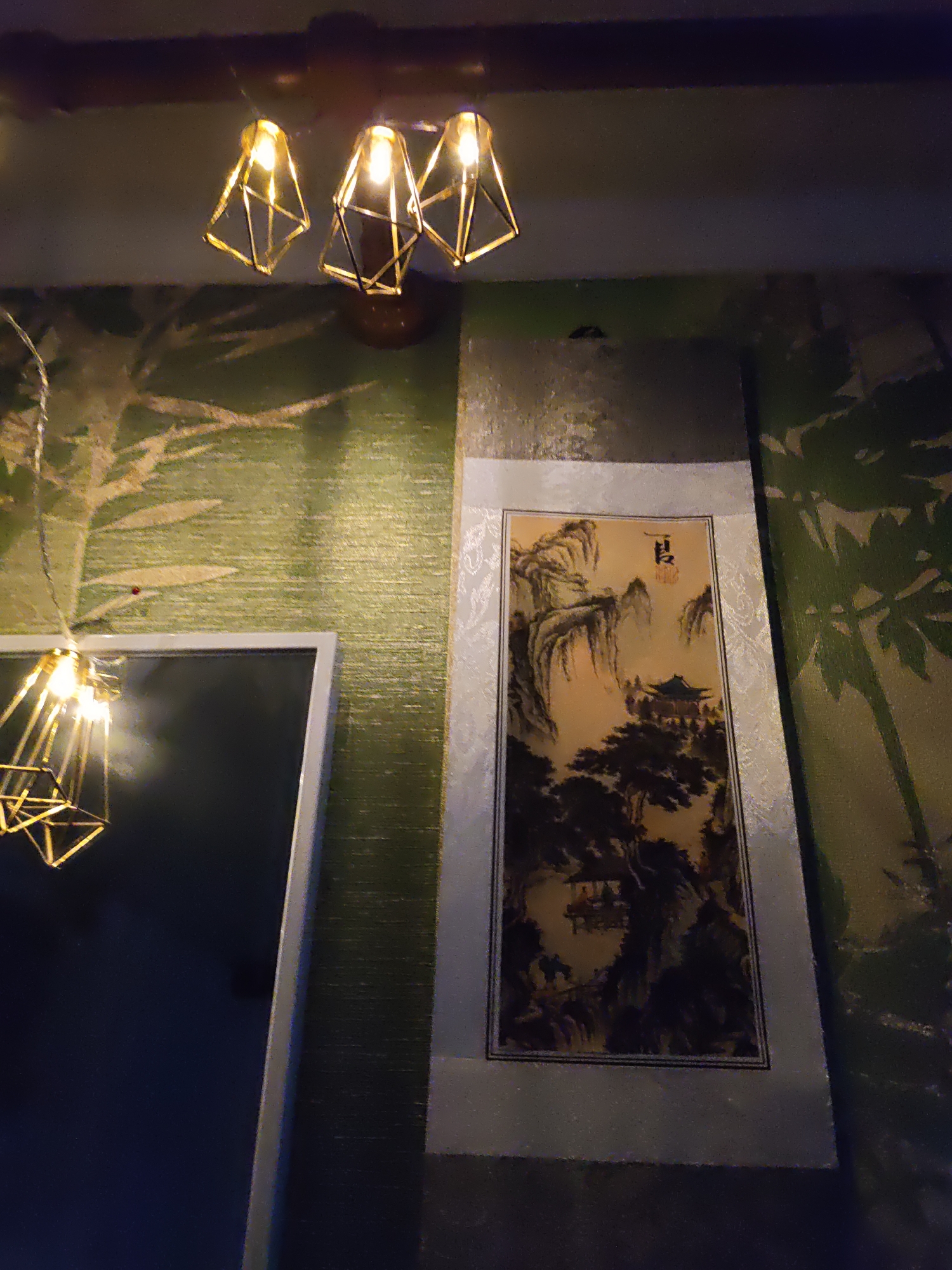 The image depicts an indoor scene featuring traditional East Asian artwork and decorative lighting. On the right side, a scroll painting displays a mountainous landscape with a building nestled amongst trees. The painting is bordered by a textured fabric. To the left, a framed mirror reflects a modern geometric pendant light fixture. Another similar light fixture is visible hanging from the ceiling above the artwork. The walls are covered in textured wallpaper, with a pattern resembling bamboo or reeds on the left side and a more abstract pattern on the right. The lighting appears to be warm and ambient, suggesting an evening or dimly lit setting. There are no people visible in the image. The overall impression is one of a tastefully decorated interior with an Asian-inspired aesthetic. This image was taken in Rostov-on-Don, Russia.