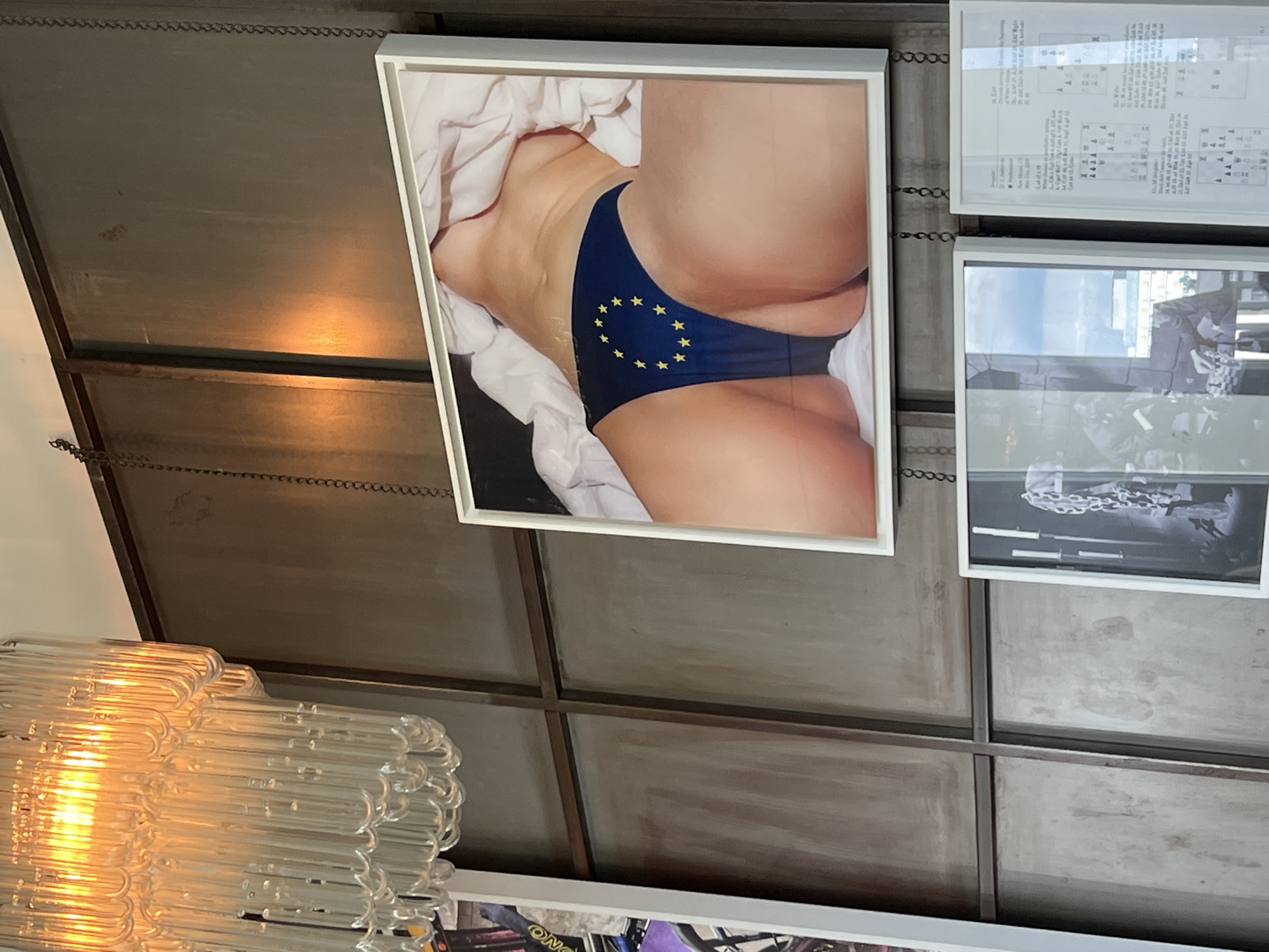 The image depicts a room with a dark, textured wall paneled with metal. A large, framed photograph hangs prominently, featuring a person's lower torso, clad in blue underwear adorned with yellow stars in a circular pattern, reminiscent of the European Union flag. The person is lying on a white surface, possibly a bed. Above this photograph, a large, multi-tiered glass chandelier with numerous cylindrical glass tubes is illuminated, casting a warm glow. To the left of the main photograph, another framed artwork is partially visible, showing a bicycle with colorful elements. Below the main photograph, two smaller framed artworks are displayed. One is a black and white photograph of what appears to be a chain or some metallic structure against a blurry background, and the other is a document with text and diagrams. The overall impression is of an art exhibition or a styled interior space, possibly in Berlin, Germany, given the contextual information provided.