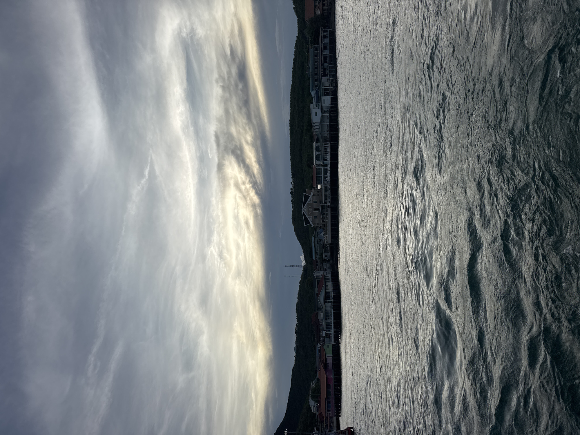 The image depicts a waterfront scene in Pattaya City, Thailand. The right side of the image shows water with rippling reflections. The left side features a coastal area with buildings nestled along the shoreline and up the hillside. The sky is filled with cloud formations.