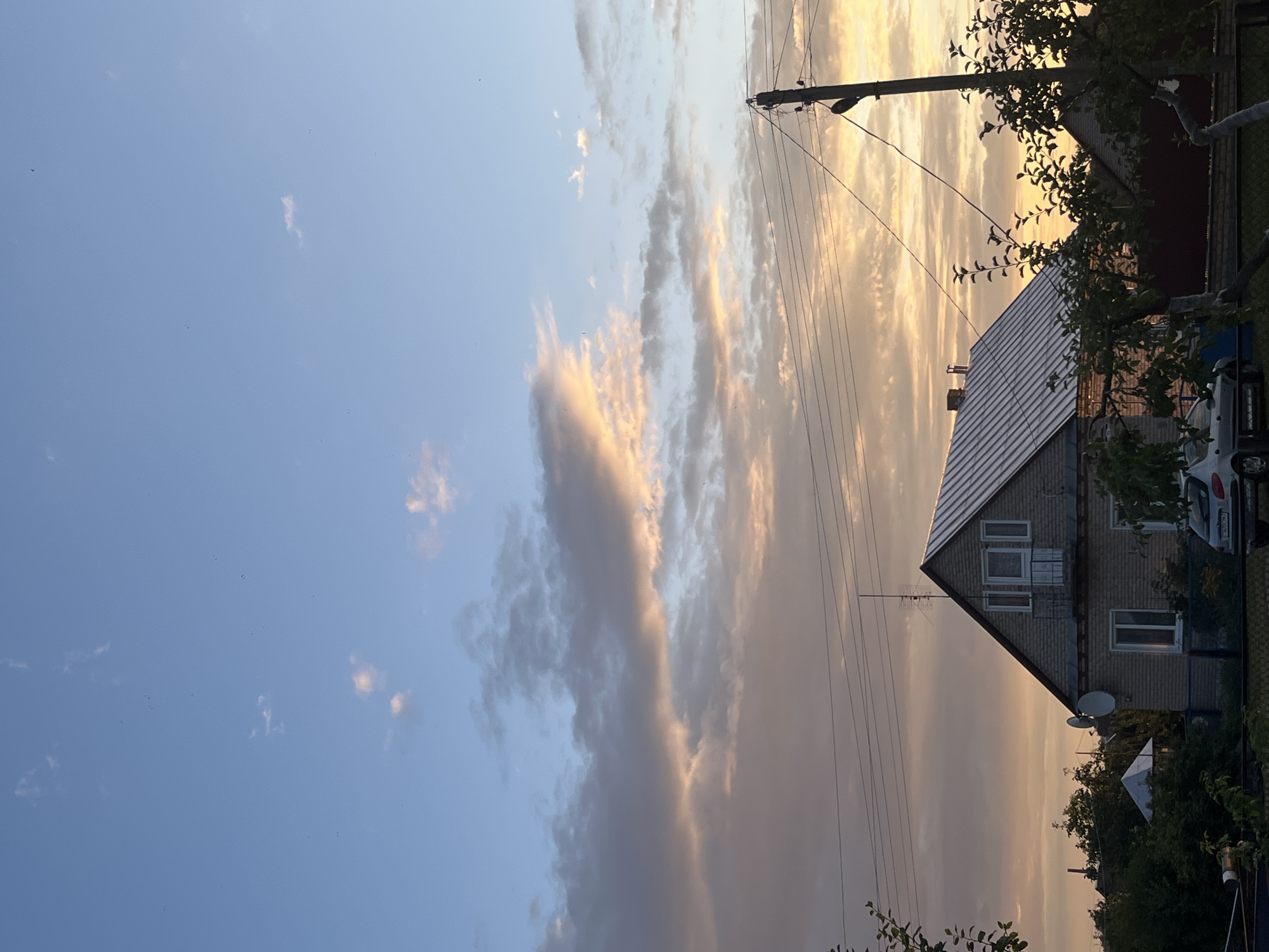 The image depicts a rural scene in the Sarootski Sel'ski Savet, Belarus, during what appears to be sunset. A two-story brick house with a silver metal roof is prominently featured, with a balcony on the upper level. Adjacent to the house, a white car is parked. Power lines traverse the sky, supported by a wooden utility pole. Lush green trees and foliage surround the property. The sky is a blend of blue, with scattered clouds tinged with warm hues of orange and pink, indicating the setting sun. The overall impression is one of a peaceful, rural dwelling at dusk.