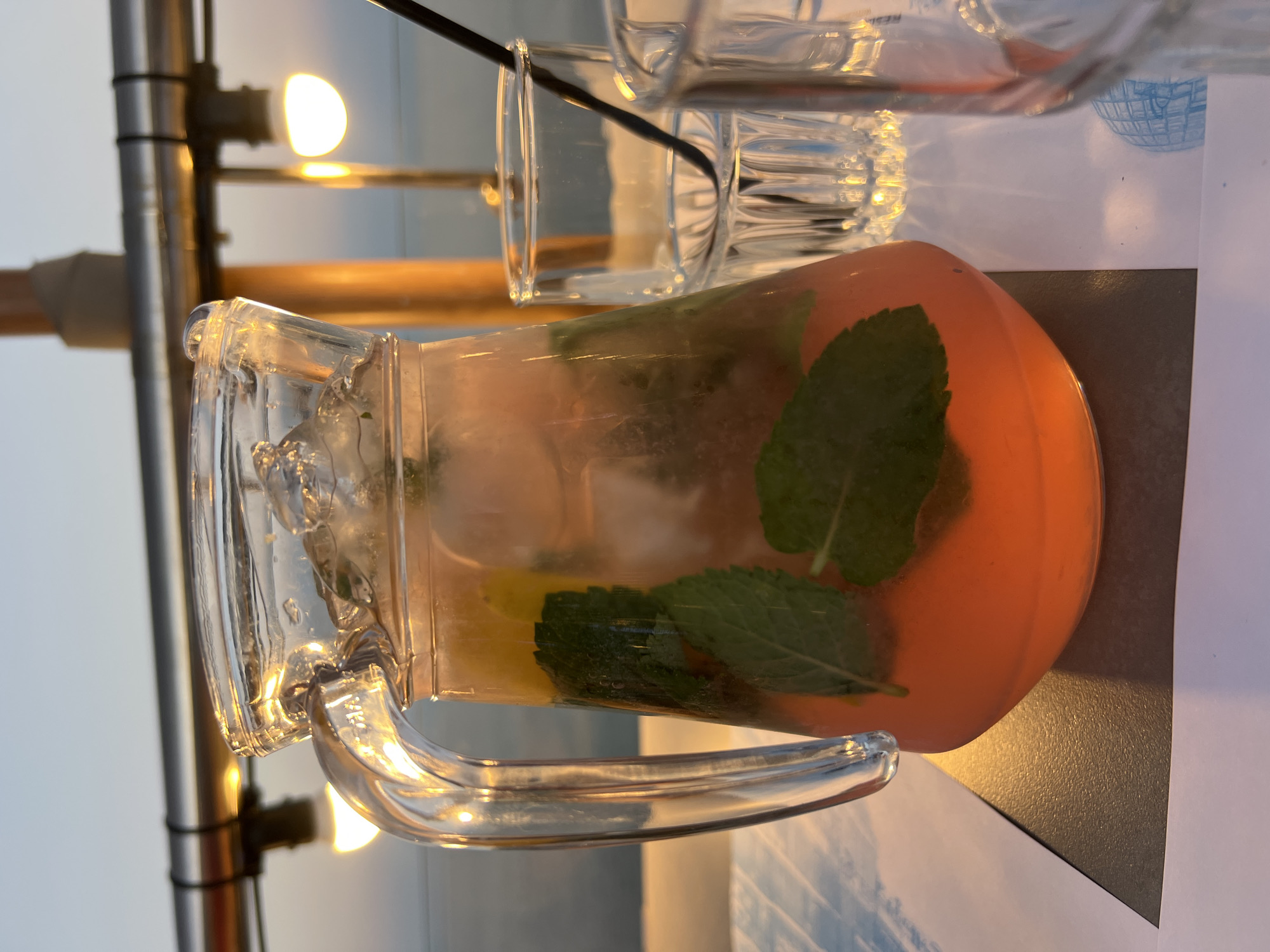 A glass pitcher filled with a pinkish-orange beverage, ice, mint leaves, and what appears to be slices of citrus fruit, rests on a table. The pitcher has a thick, clear glass handle. Next to the pitcher are two empty, clear glass tumblers, one of which has a black straw inserted. The table is covered with a light-colored, possibly paper, tablecloth with a blue design. In the background, a metal railing with lights is visible, casting a warm glow. The lighting suggests it is either dawn or dusk. The overall scene appears to be an outdoor seating area of a restaurant or cafe, possibly with a view of water, given the hazy horizon. The location is Odesa, Ukraine.