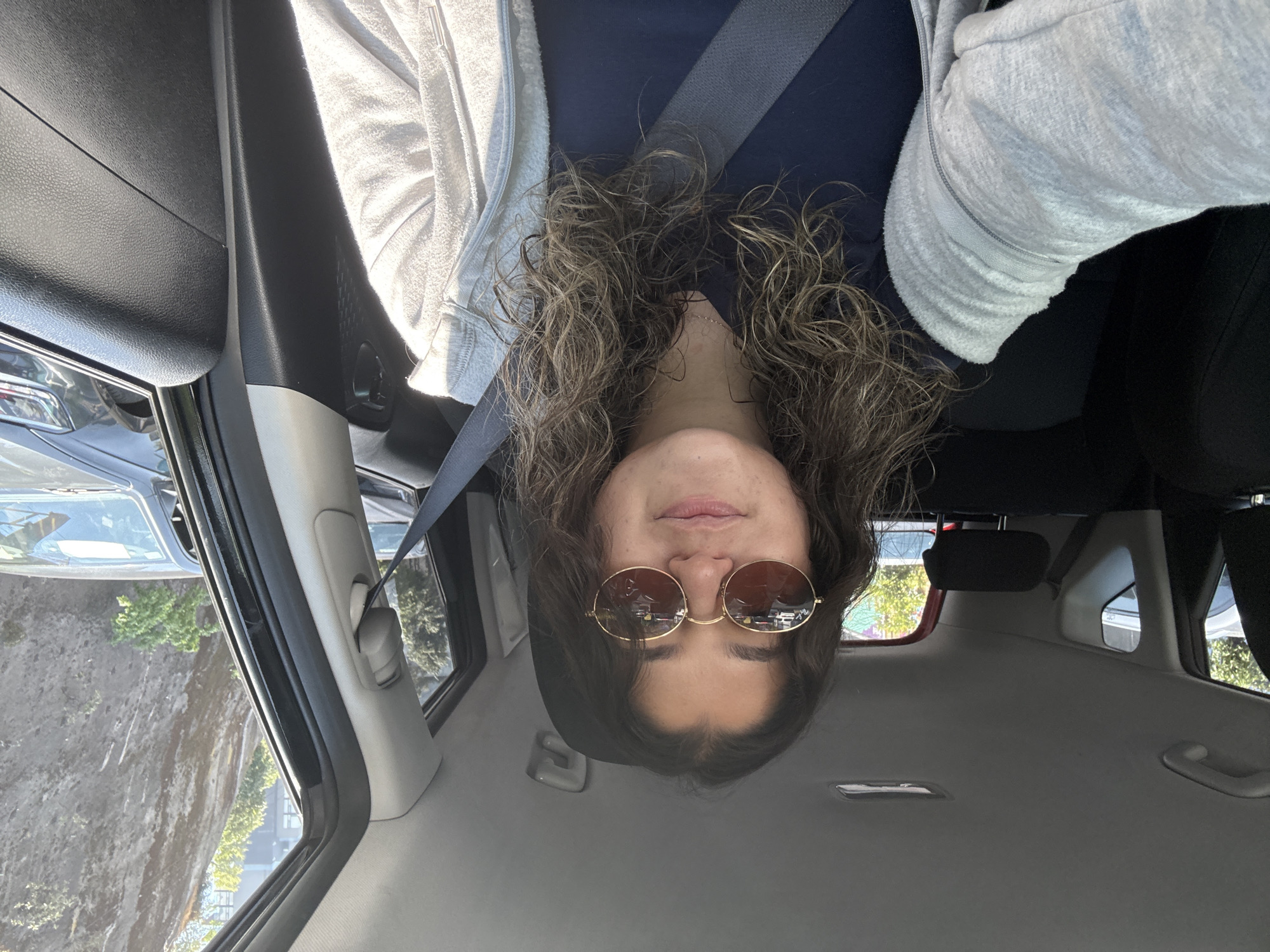 An inverted portrait-style image captures a person from a low angle within a car. The subject, a person with medium-length, curly brown hair, is looking downwards, with their head positioned at the bottom of the frame. They are wearing round, gold-rimmed sunglasses that reflect the exterior environment. Their lips are slightly parted, and their chin is visible. The person is dressed in a dark blue top and a light gray, textured, zippered hoodie, over which a gray seatbelt crosses their chest.
The setting is the interior of a car, with elements like the dark gray dashboard or door panel visible on the left, a gray car ceiling, and parts of the car's seating structure. Through the car windows, bright daylight illuminates the scene. On the left side of the image, outside the car, a large, light-colored rock formation or cliff face with sparse green vegetation is clearly visible under sunny conditions. Reflections in the sunglasses hint at a cityscape with buildings and trees. No discernible text is visible within the image. The overall impression is a casual, sunlit moment inside a vehicle, possibly in an area surrounding Mexico City, Mexico, given the diverse landscape.