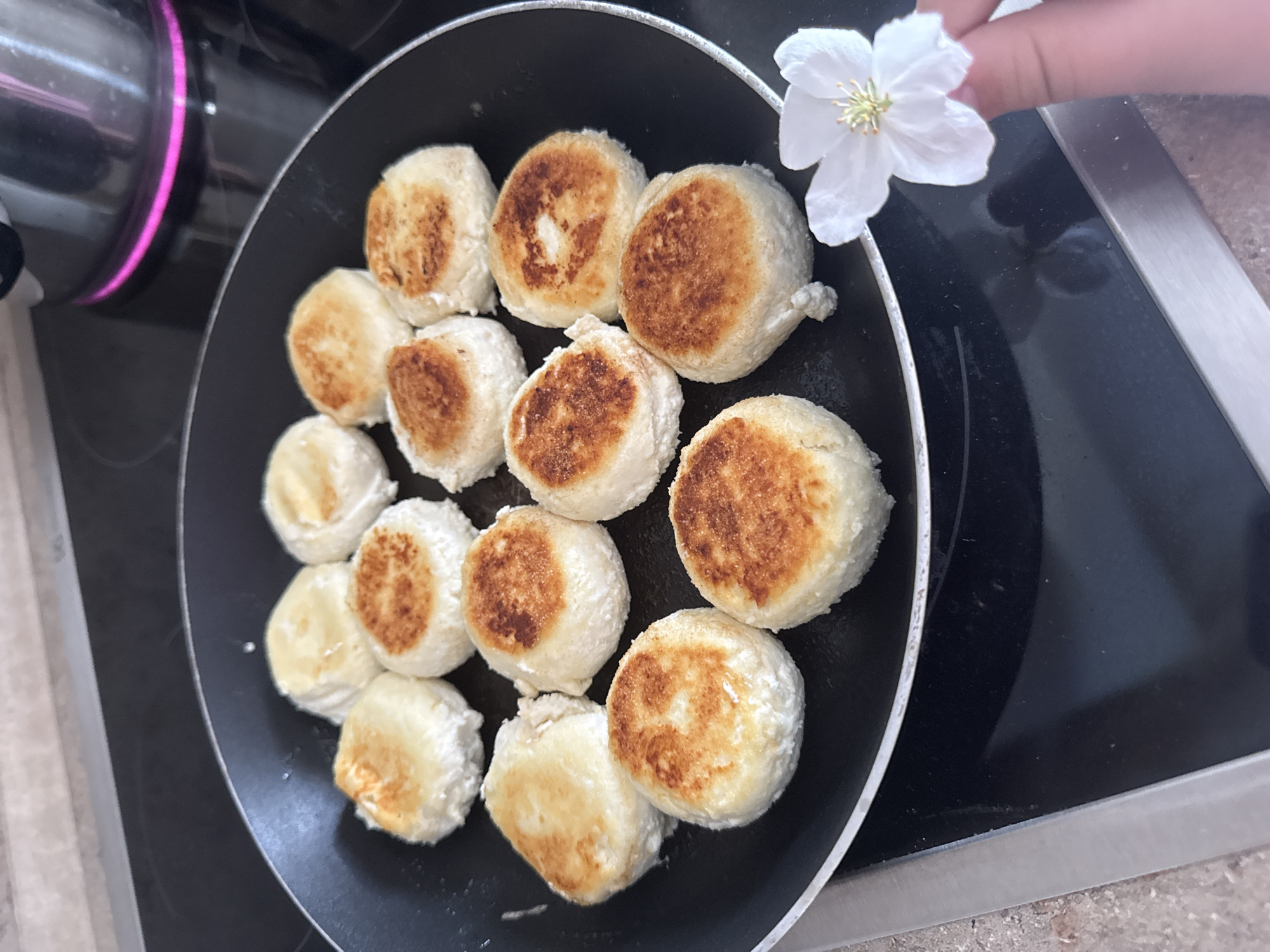 A high-angle shot shows a black frying pan filled with about 14 small, round cheese pancakes, some browned on one side, sitting on a cooktop. A person's hand holds a white flower near the pan's edge. The cooktop is stainless steel and black. The image does not provide any identifiable information about Dzerzhinsk, Russia.