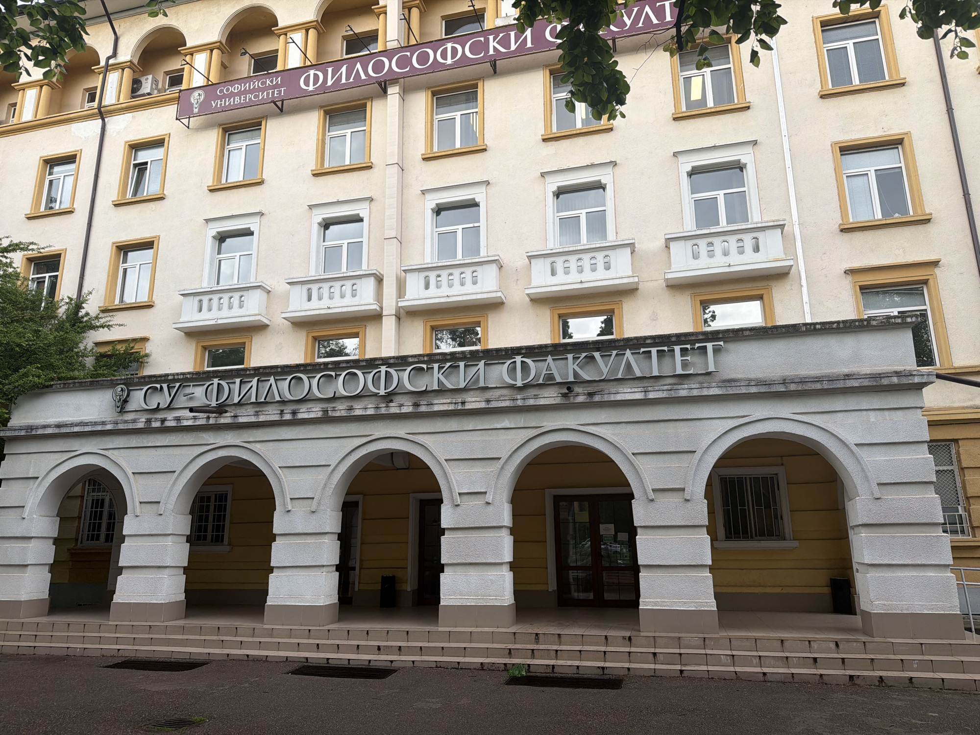 The image shows the facade of the Faculty of Philosophy at Sofia University "St. Kliment Ohridski" in Sofia, Bulgaria. The building is light yellow with white architectural details, including arched doorways and window frames. A sign above the entrance reads "СУ - ФИЛОСОФСКИ ФАКУЛТЕТ" (SU - Faculty of Philosophy) in Bulgarian. Above the second story is another sign that reads "СОФИЙСКИ УНИВЕРСИТЕТ ФИЛОСОФСКИ ФАКУЛТЕТ" (Sofia University Faculty of Philosophy) in Bulgarian. The building appears to be a multi-story structure with rows of windows. The foreground shows a set of steps leading up to the entrance. The sky is overcast, and some greenery is visible on the left side of the frame. There are no people visible in the image.
