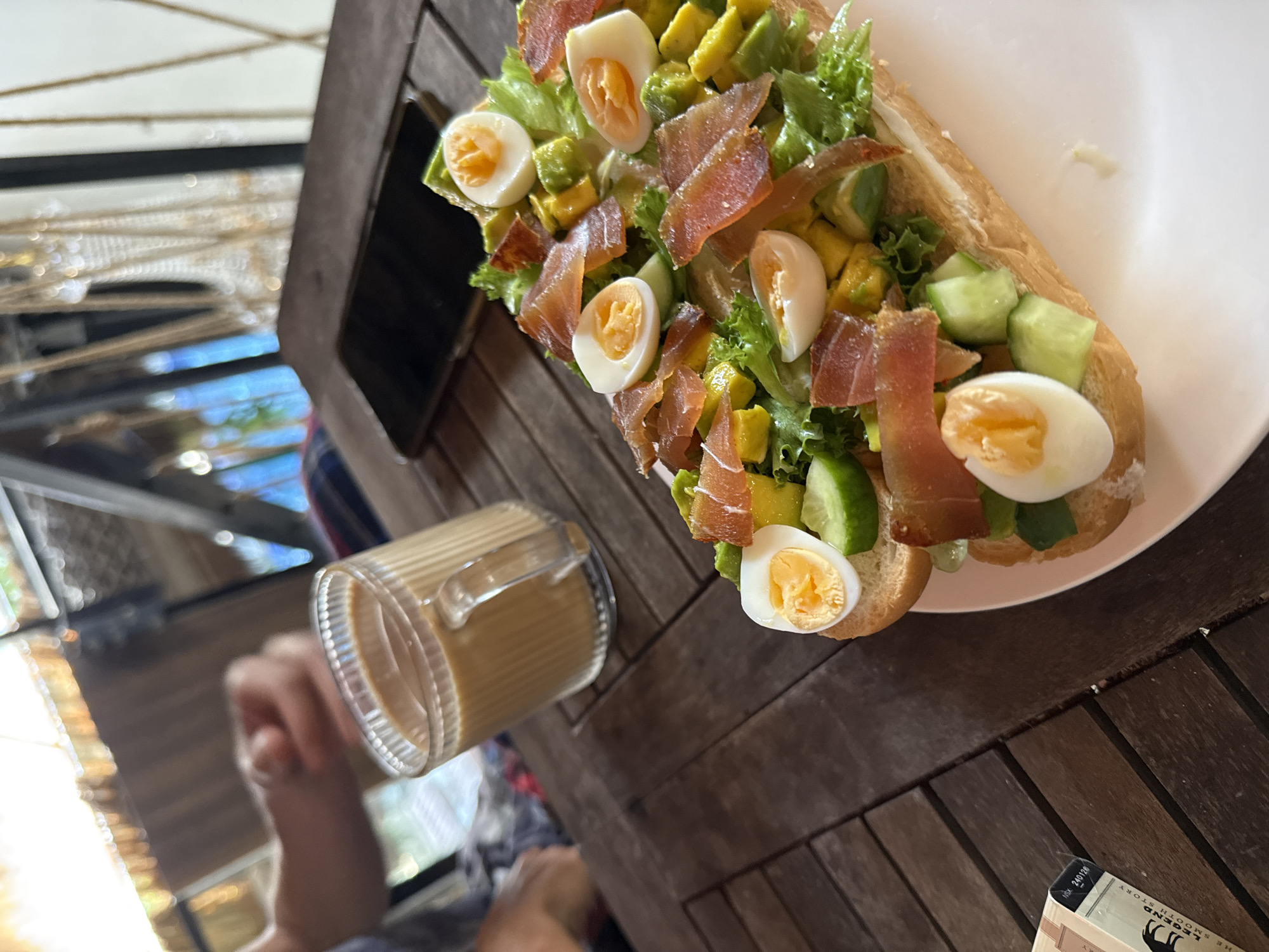 A close-up, angled shot captures a meal and drink on a wooden table in what appears to be a restaurant setting in Bac Natural Heritage Area Trang, Vietnam. A sandwich, topped with lettuce, avocado, boiled egg slices, smoked fish, and cucumber, sits on a white plate. To the left, a glass mug containing a light brown beverage is visible. A hand holding the glass is partially visible. A black smartphone is partially visible. The overall tone suggests a casual dining experience.