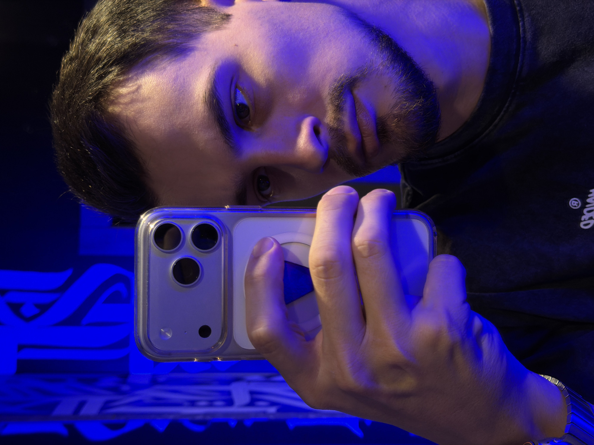 A close-up, tilted shot shows a person holding a smartphone up towards their face. The person has dark hair, a beard, and is wearing a dark shirt with some text visible on the sleeve. The smartphone is in a clear case and features three camera lenses and a pop socket. The scene is bathed in blue light. The background shows a blue patterned surface. It is not possible to determine the exact location in Krasnoyarsk, Russia based on the provided image.