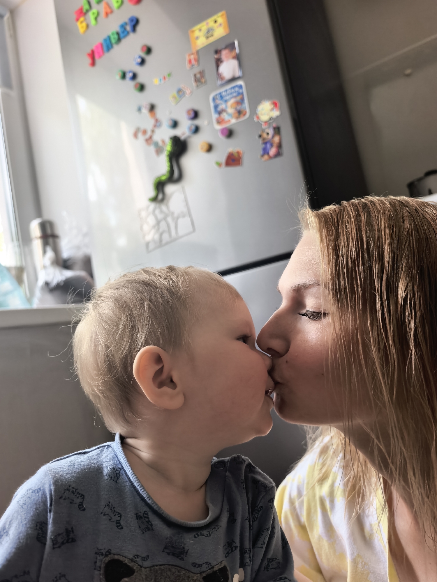 A heartwarming, close-up vertical photograph captures a tender moment between a woman and a young child indoors in Sterlitamak, Russia. The two subjects are positioned in profile, their noses touching as they share a gentle kiss. The woman, with long, light brown hair that appears slightly damp, has her eyes closed in an expression of deep affection. She is wearing a yellow and white patterned top. The child, likely a toddler, has light blonde hair and is dressed in a blue long-sleeved shirt adorned with small animal patterns, possibly raccoons. The child's eyes are open, looking towards the woman.
The background indicates a domestic setting, likely a kitchen or a similar area. A large, light-colored refrigerator, possibly stainless steel, is prominently featured, decorated with numerous colorful magnetic letters and various picture magnets, including personal photos and cartoon characters. A distinct green, snake-like or lizard-shaped magnet is also visible on the fridge. Among the magnetic letters, Cyrillic words such as "МАМА" (Mama), "БРАВО" (Bravo), and "УМНИЦА" (Umnitsa, meaning "clever girl" or "well done") are discernible. To the left, a window allows bright natural light to illuminate a window sill where a silver thermos and other indistinct items are placed. The scene evokes feelings of love and warmth within a cozy home environment during the daytime.