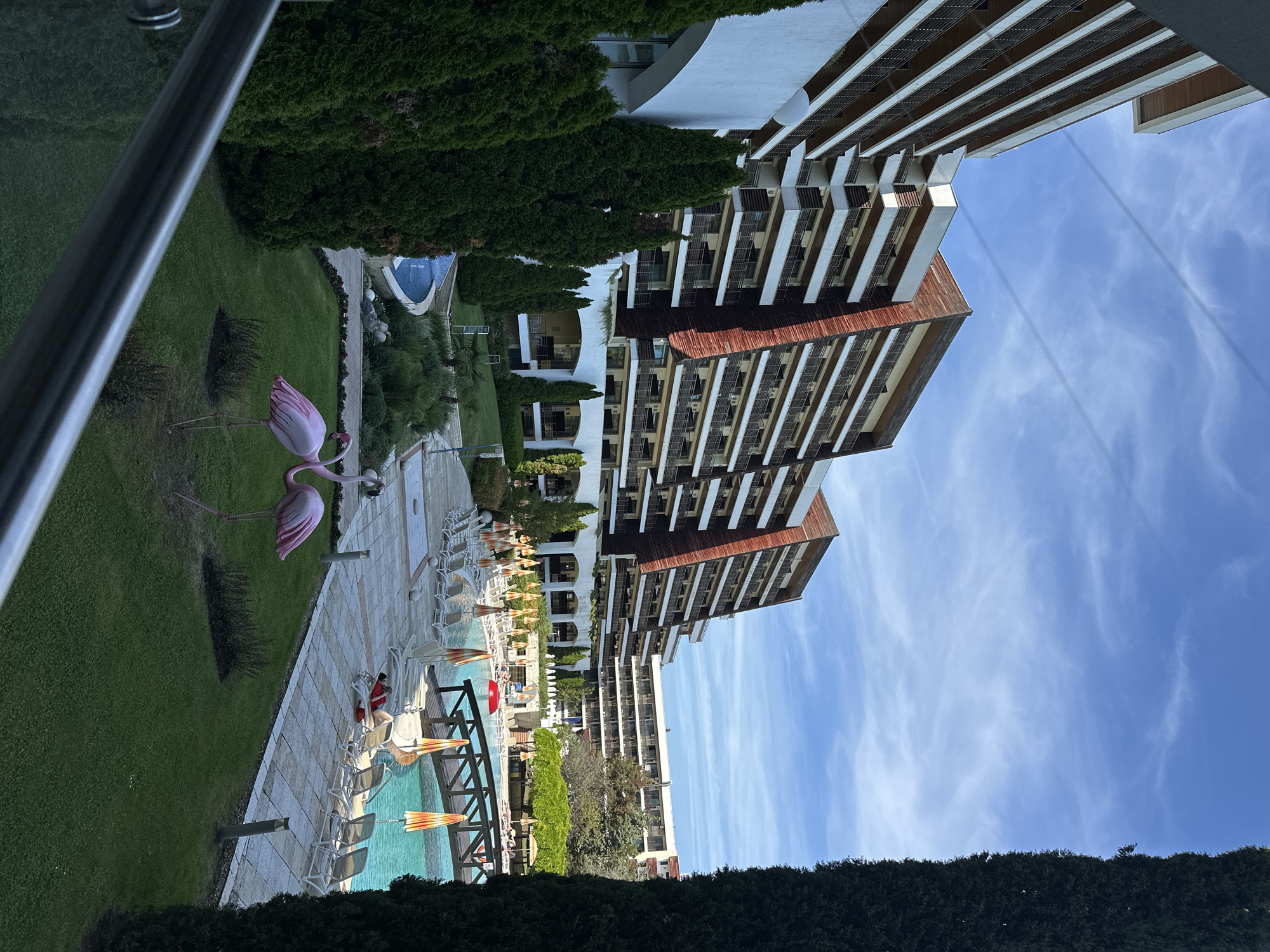 This media file captures an outdoor scene at a resort or hotel, identified as being in Obrochishte, Bulgaria. The image is taken from an elevated perspective, likely a balcony, looking down and across the property.
The primary subjects include several large, modern multi-story buildings, presumably hotels or apartment blocks, with numerous balconies and a facade featuring white and brown/wood-like elements. In the foreground, a metal railing is visible on the left, and a dark green hedge extends across the bottom.
The central part of the scene is dominated by a expansive swimming pool area. The light blue water of the pool is surrounded by a paved patio lined with numerous white sun loungers and some striped orange-and-white parasols. A wooden bridge or walkway crosses a section of the pool. One person in a red top is visible sitting on a sun lounger by the pool.
Adjacent to the pool area is a well-maintained green lawn where two prominent pink flamingo statues stand. Dense green hedges and various trees frame the property and separate different sections of the landscape. In the distance, another, smaller pool with a distinct shape is visible nestled among greenery.
The sky is clear and bright blue with scattered thin, white clouds, indicating pleasant, sunny weather. The bright light and relatively short shadows suggest it is daytime, likely late morning or early afternoon. No visible text is discernible in the image. The overall atmosphere conveys a sense of leisure and vacation at a resort property.