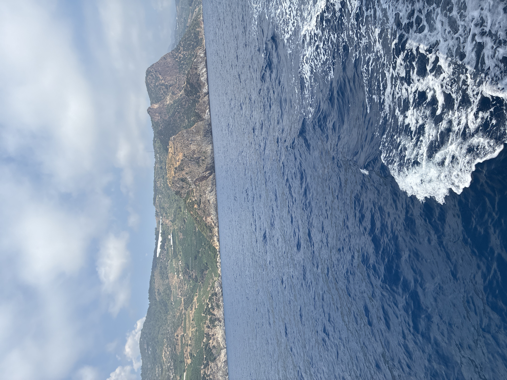 The image depicts a view of the Mediterranean Sea from a moving boat, with the rugged coastline of Gazipaşa, Turkey, in the background. The sea is a deep blue, with white foam trailing behind the boat in the foreground. The coastline is characterized by steep, rocky hillsides covered in sparse vegetation, with some areas appearing to be cultivated with terraces. White structures, possibly greenhouses or buildings, are visible on some of the hillsides. The sky is partly cloudy, with white cumulus clouds against a pale blue background. The overall impression is one of a sunny day at sea along a dramatic coastal landscape.