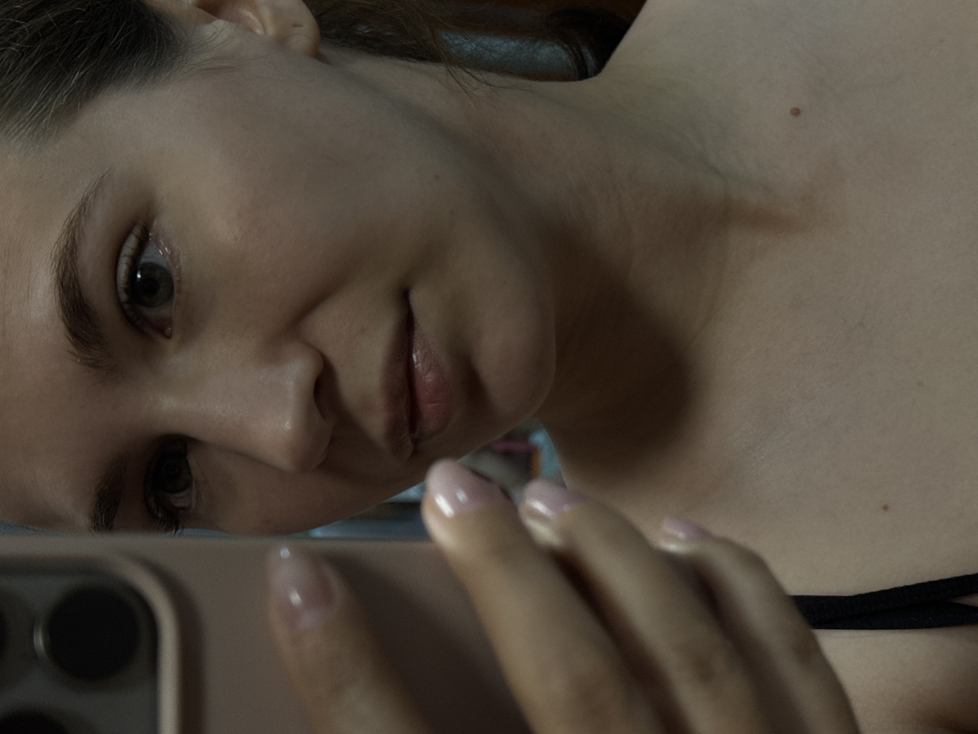 The image shows a close-up of a young woman taking a selfie with a smartphone. Her left hand, with manicured nails, is holding the phone. Her face is partially visible, with her eye, nose, and lips in focus. She is wearing a black top. The background is blurred, but appears to be an indoor setting. There is no information in the image to suggest a location in Chaikovsky, Russia.