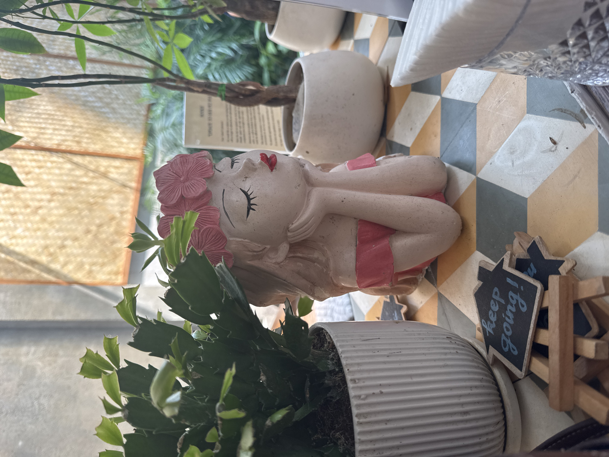 The image depicts a tabletop scene, likely indoors, in Đà Nẵng, Vietnam, featuring a decorative figurine of a woman with red lips and a flower crown, resting beside a small chalkboard sign that reads "keep going!". The figurine is positioned next to a white ribbed planter containing a green succulent-like plant. Another white pot with a tree trunk is present in the background. The surface underneath features a geometric tiled pattern in colors of white, gray, and gold. Other unidentifiable objects are partly visible in the background and corners.