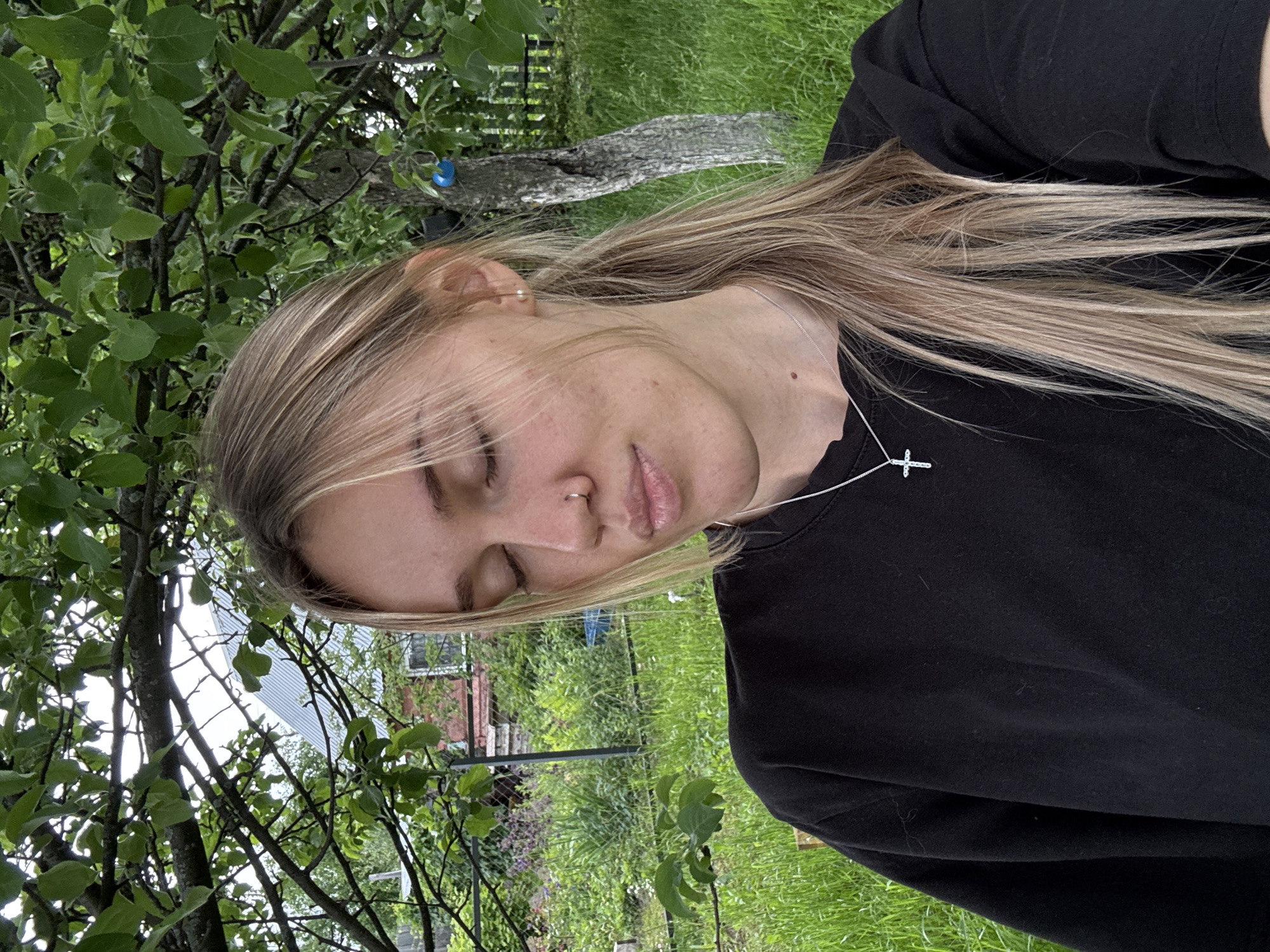 A close-up outdoor portrait features a young Caucasian female, set in Chaikovsky, Russia. She has blonde hair, with strands framing her face and a ponytail visible on her right. Her eyes are closed, and her lips are slightly pursed. She wears a silver stud nose piercing on her right nostril and a silver chain earring with a pearl in her right ear. A silver cross pendant necklace is worn over a black long-sleeved top. A small mole is visible on her left clavicle area. The background is dominated by lush green foliage, including trees and grass. A red brick house with a white window is partially visible through the foliage on the left, with a metallic corrugated roof of a building appearing behind it. A wooden fence is also visible in the background. A small blue decorative object hangs from a tree branch on the right. The scene is lit by diffused light, indicating an overcast sky, within a verdant outdoor space, likely a garden or rural yard.